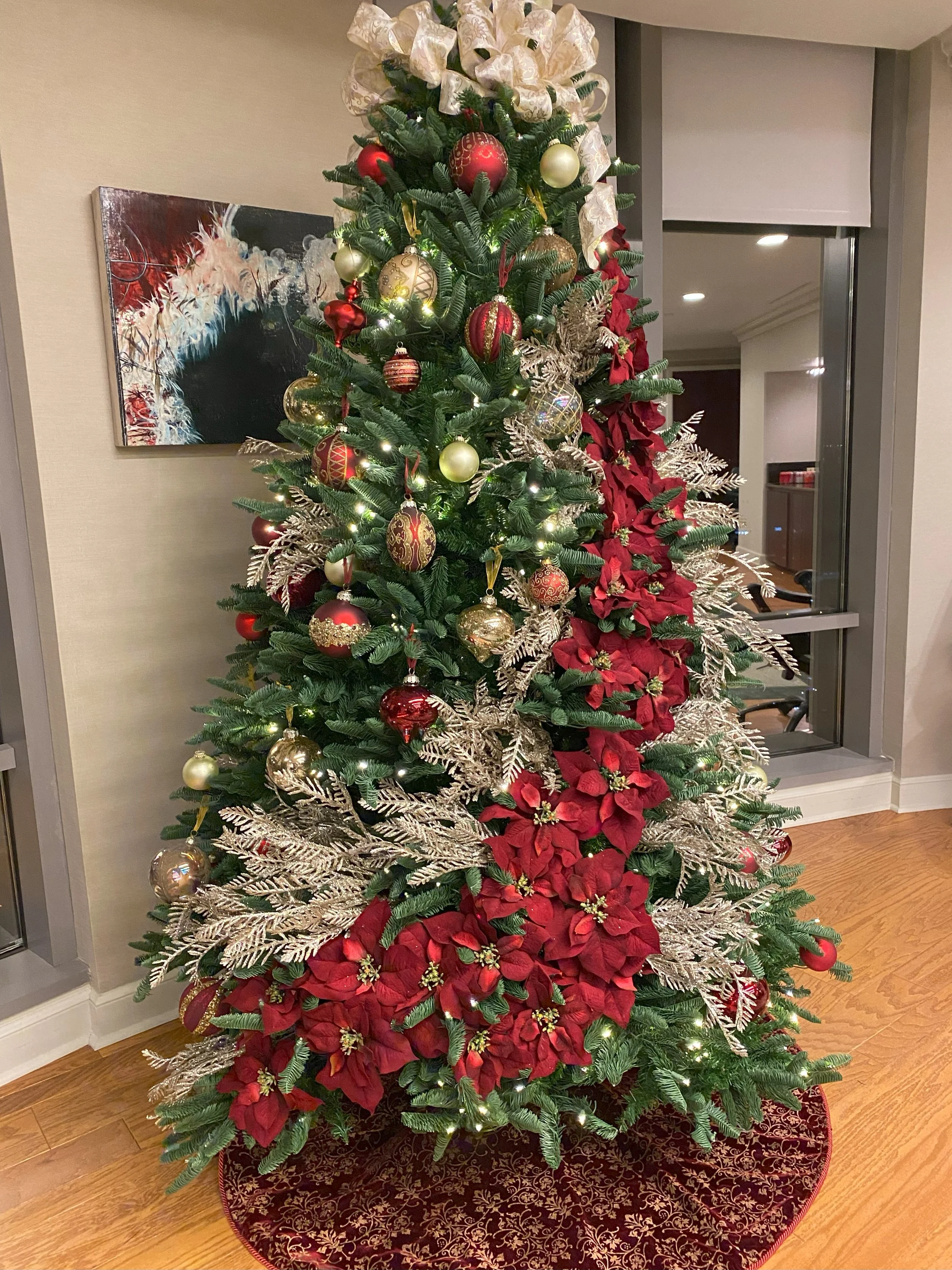 Decorated Christmas tree with red, gold, and green ornaments, gold and red poinsettias, and gold accents, sitting on a red and gold patterned tree skirt.