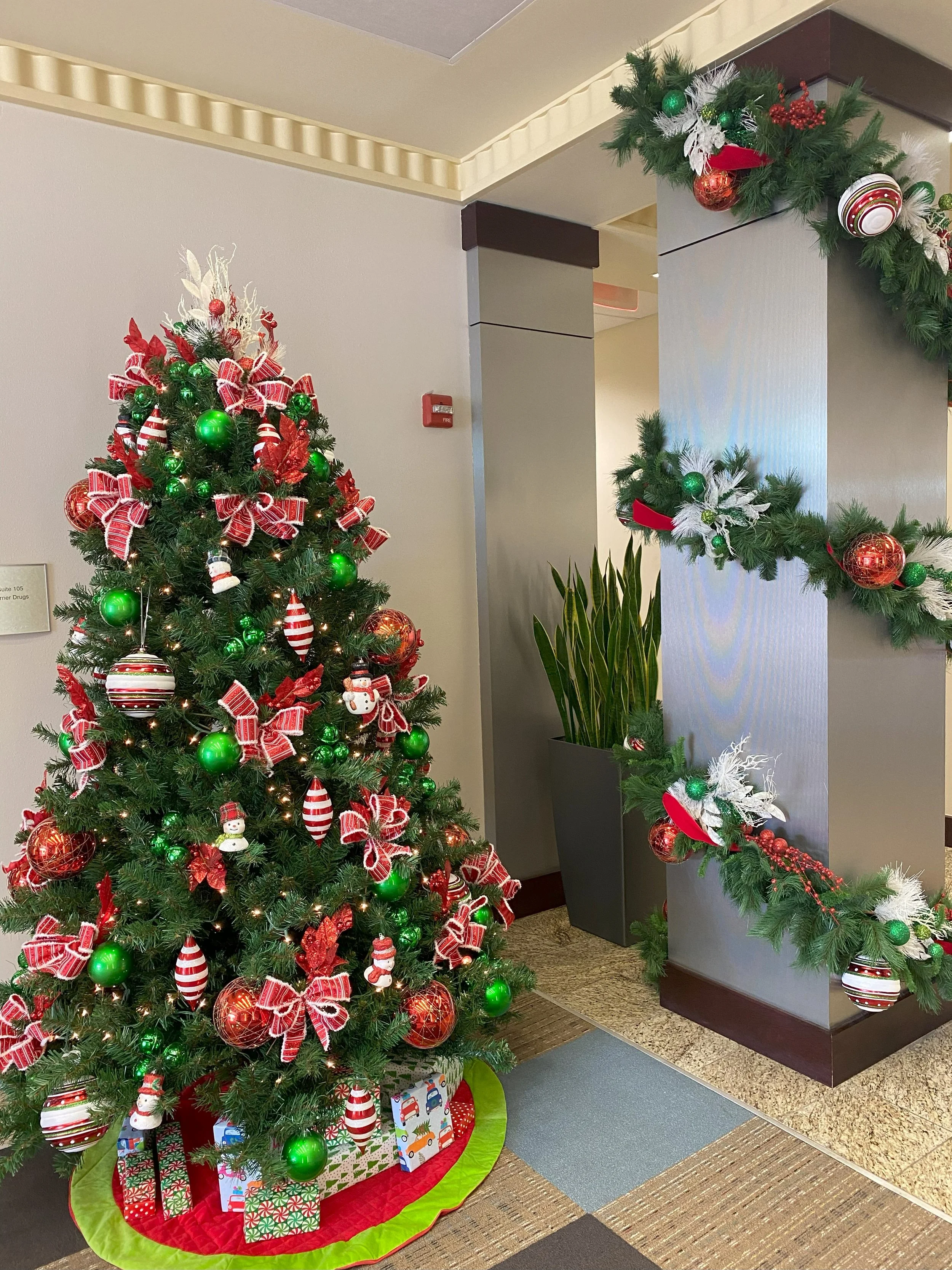 Christmas decorated tree with red and green ornaments, ribbons, and presents underneath, next to a gray wall with holiday garland.
