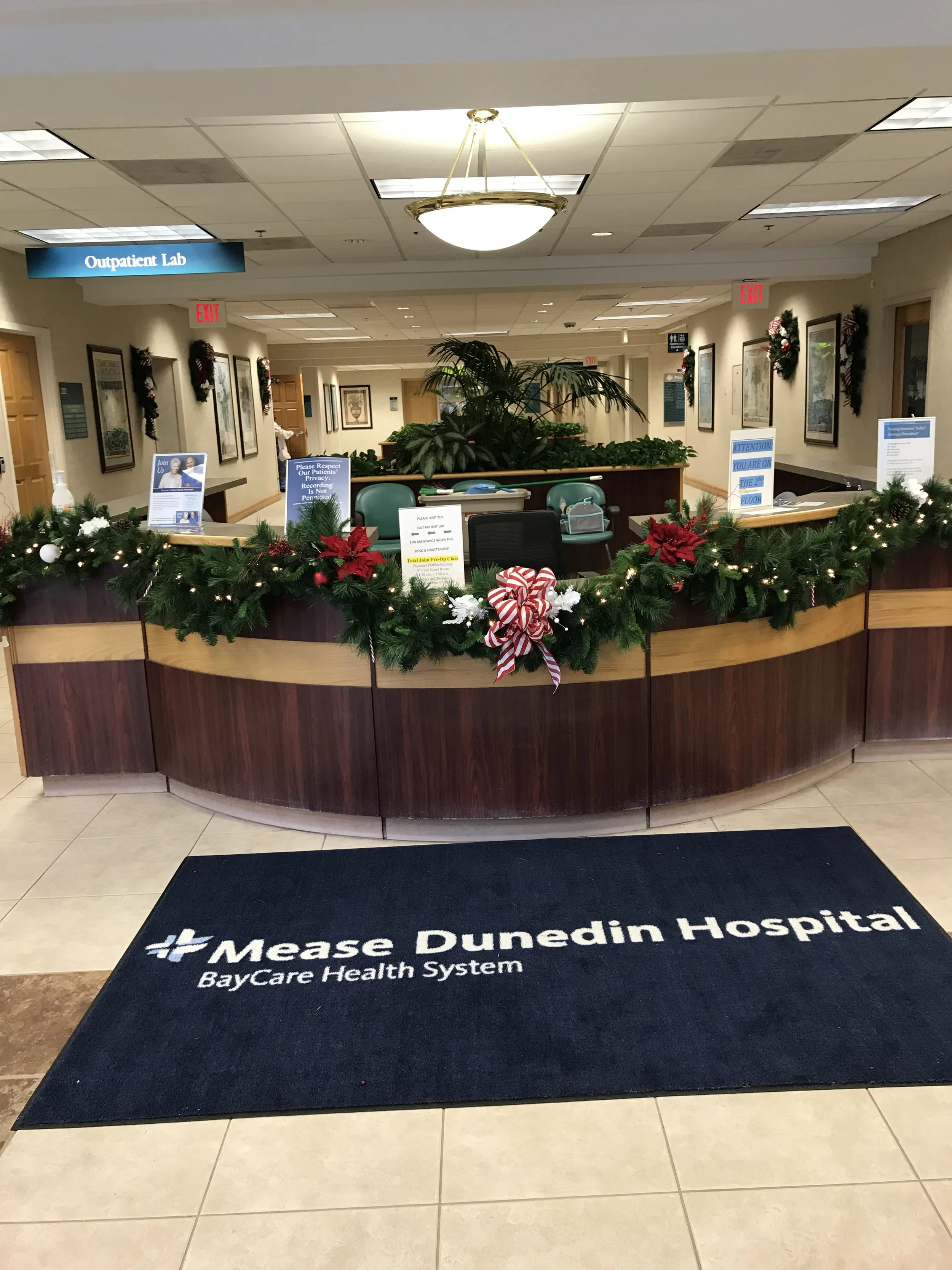 Reception area at Mease Dunedin Hospital decorated with holiday garlands and ornaments, including red poinsettias, white baubles, and a large red-and-white striped bow, with signs and plants behind the counter.