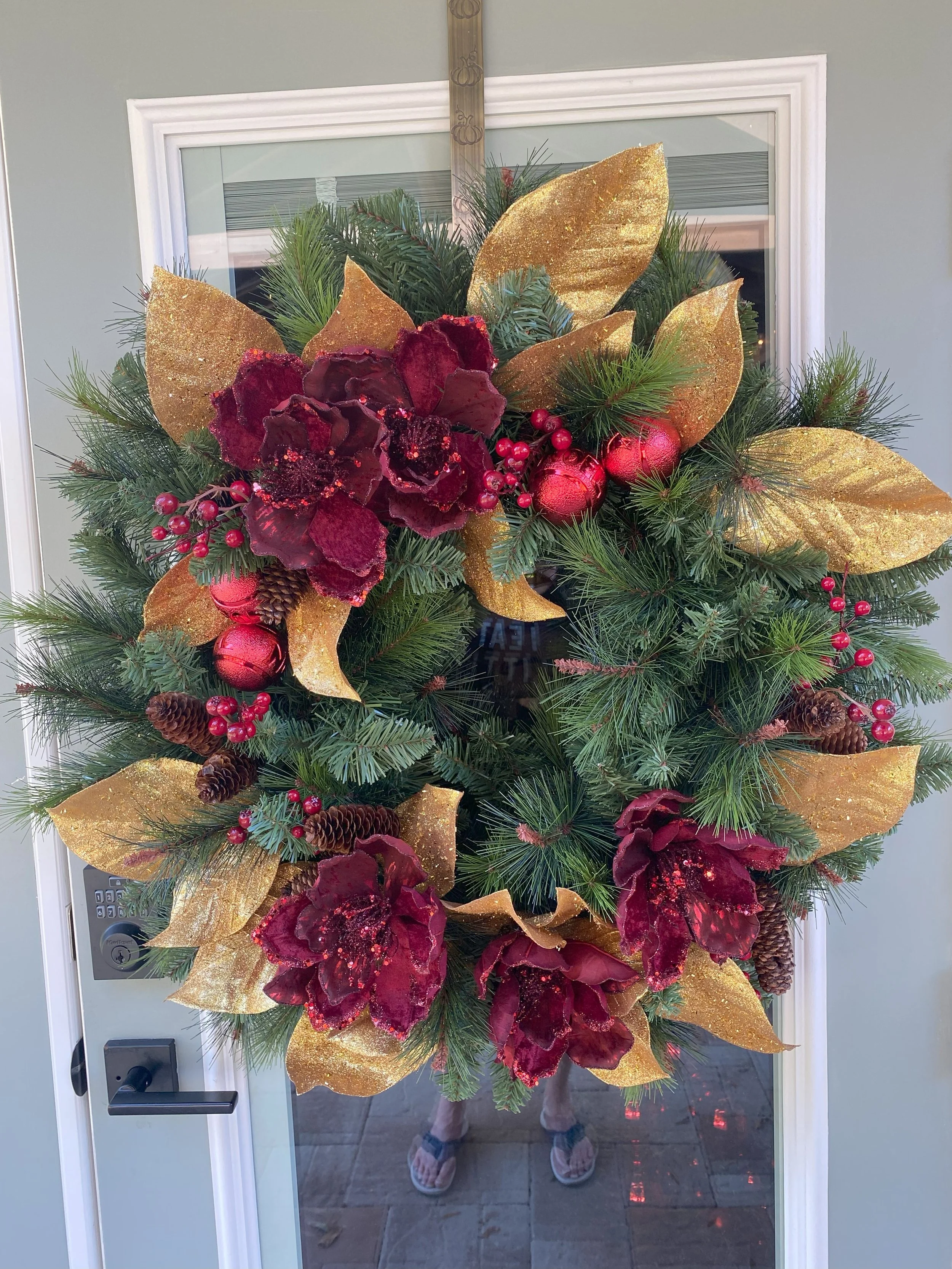 Decorative Christmas wreath on a glass door with red flowers, gold leaves, pinecones, red berries, and Christmas ornaments.