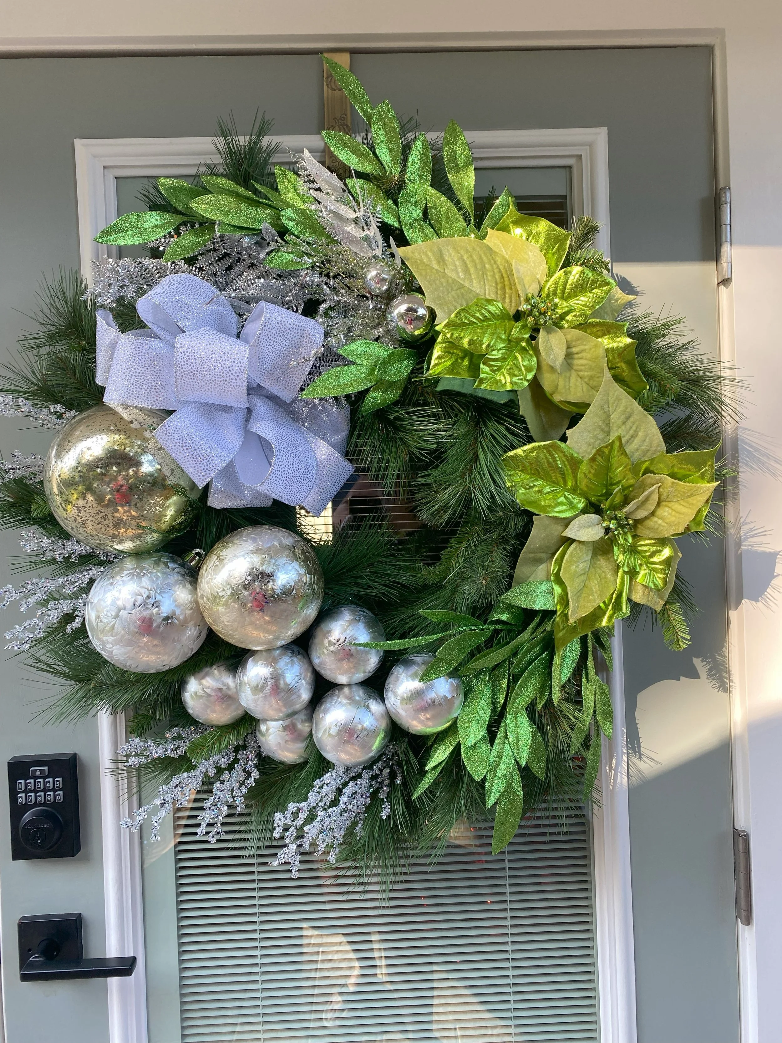 A decorated Christmas wreath with silver ornaments, a large white bow, and green foliage hanging on a door.