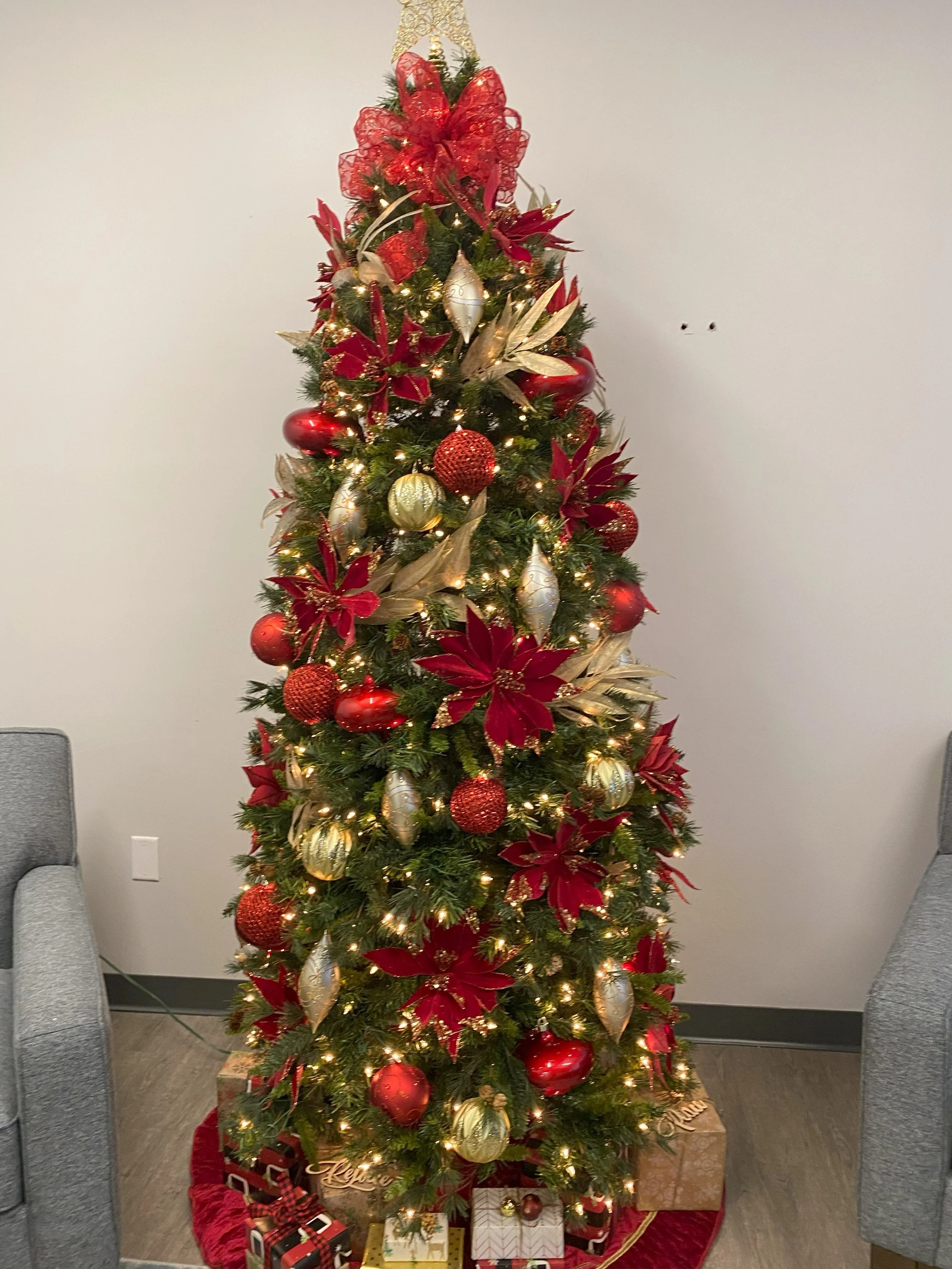 Decorated Christmas tree with red, gold, and white ornaments, poinsettia ornaments, and string lights, situated indoors on a red tree skirt with wrapped gift boxes underneath.