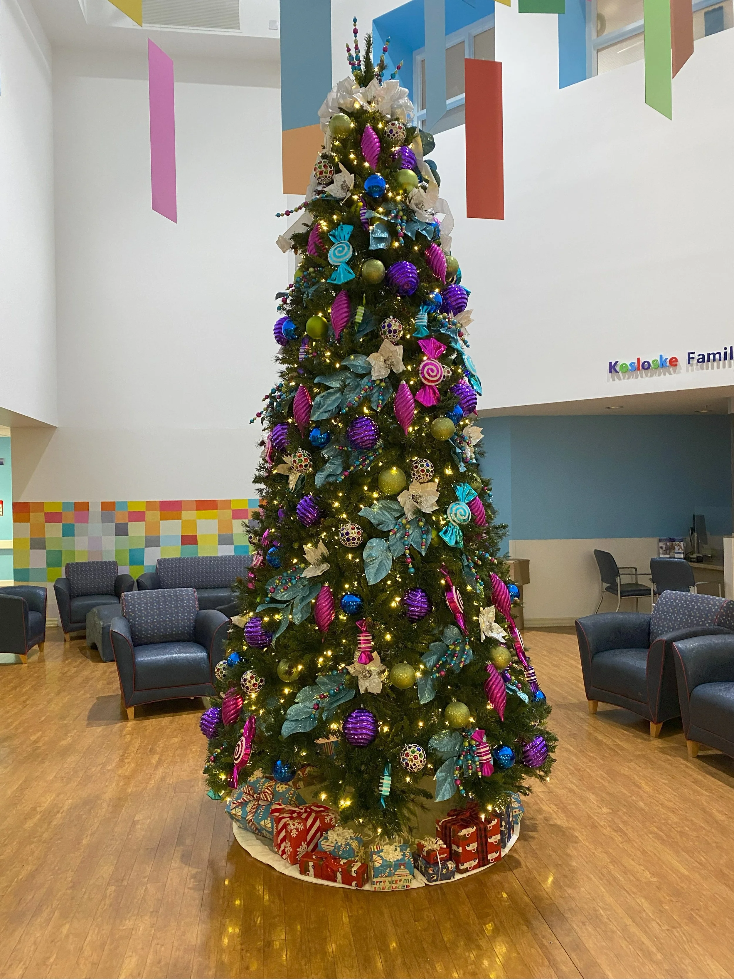 A decorated Christmas tree with purple, gold, and blue ornaments, surrounded by wrapped presents at the base, in a festive indoor setting.