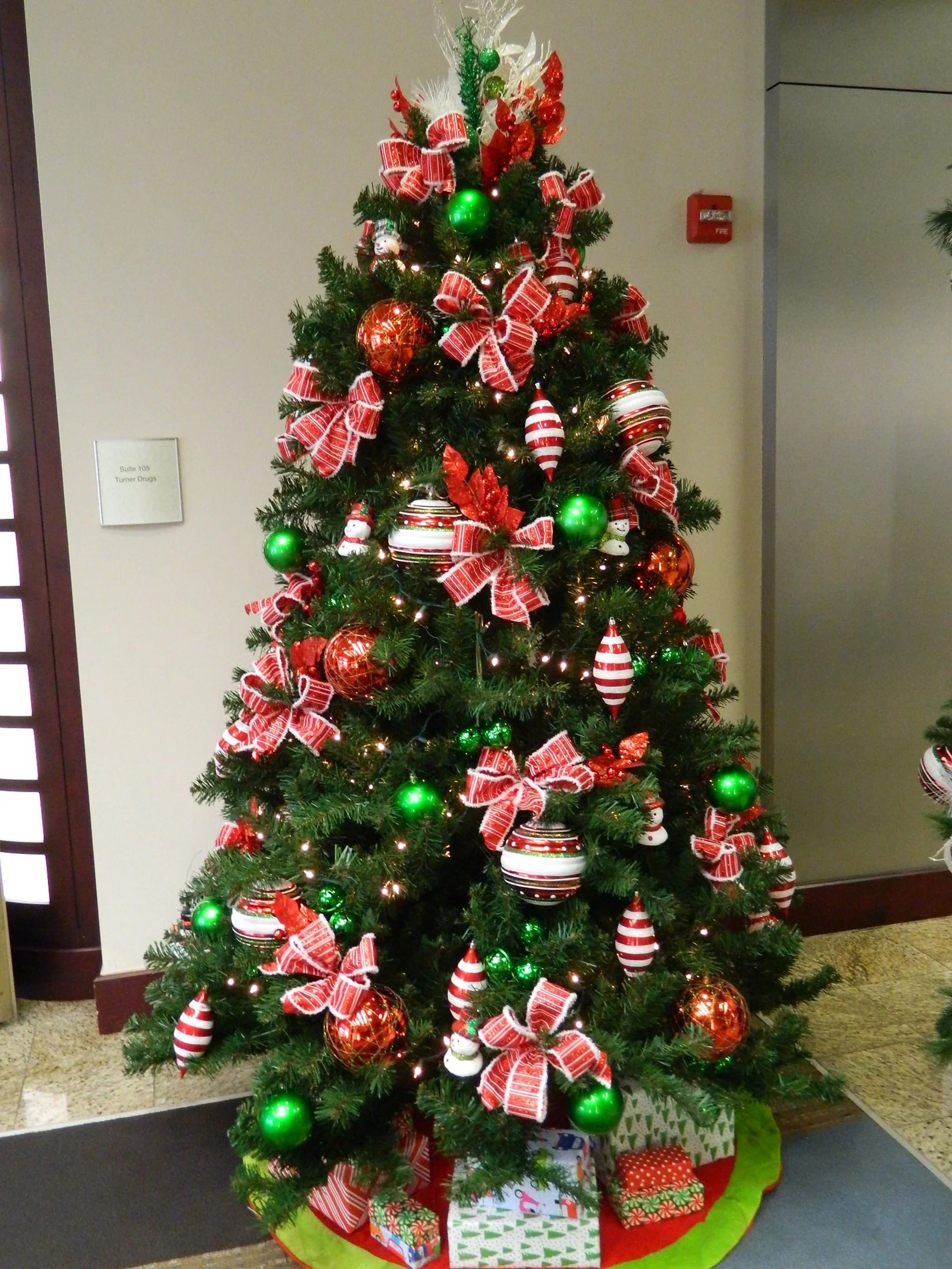 Christmas tree decorated with red, green, and white ornaments, ribbons, and lights, with wrapped gifts underneath.