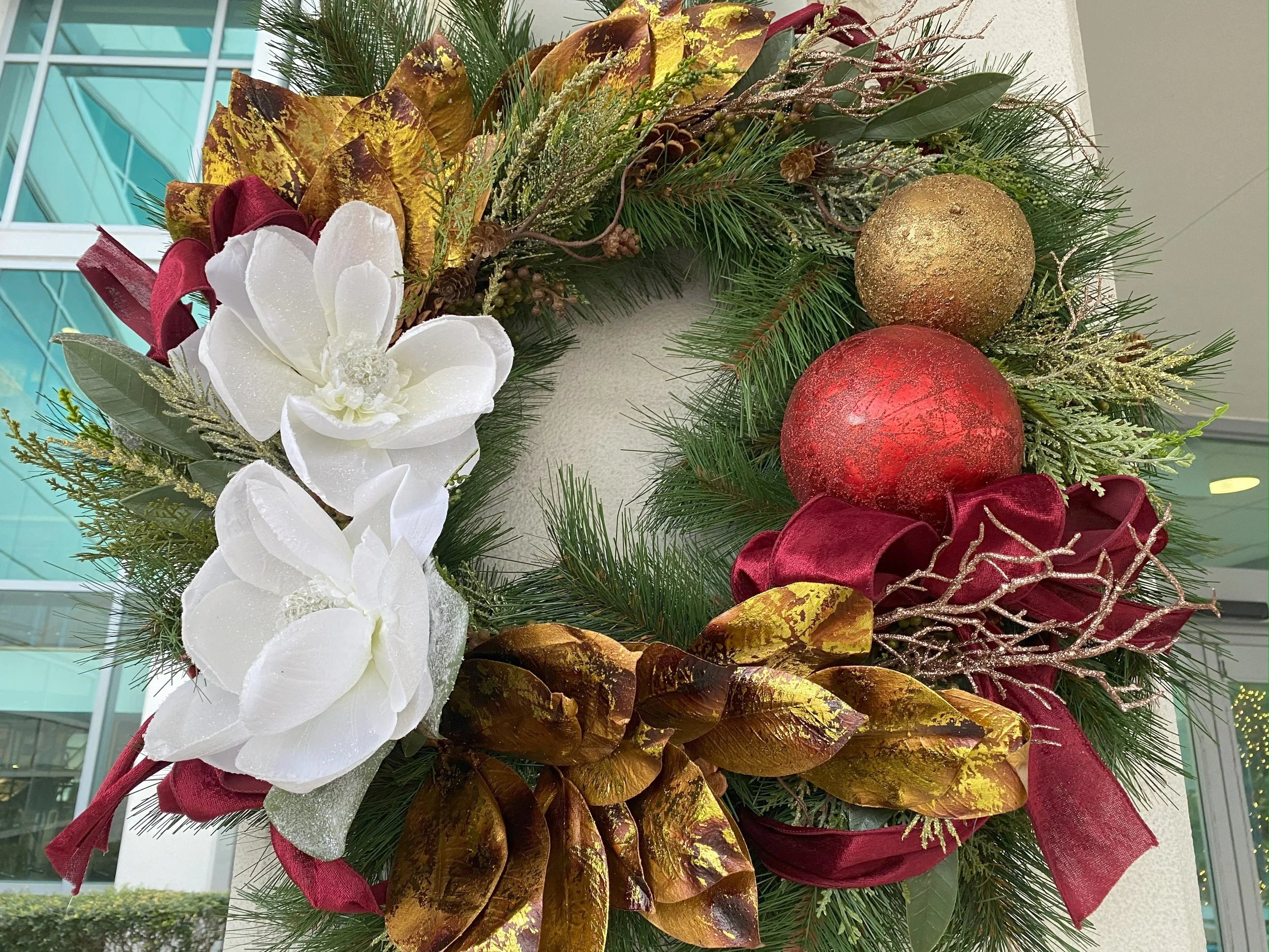 Christmas wreath decorated with white flowers, red and gold ornaments, red ribbon, gold and green leaves, pine branches, and gold glitter accents.