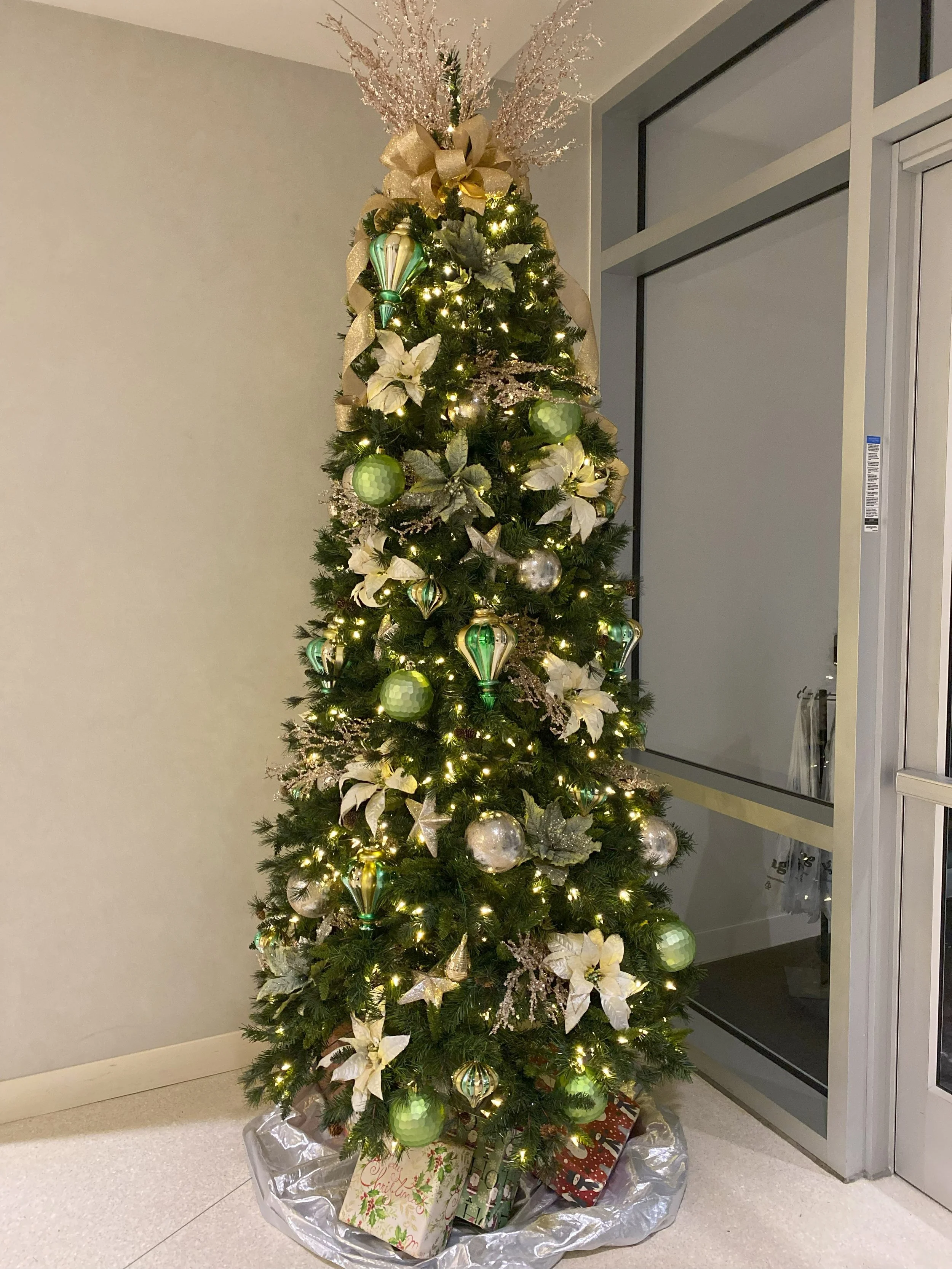 Decorated Christmas tree with white poinsettias, green and silver ornaments, gold ribbon, and white lights, with wrapped gifts underneath.