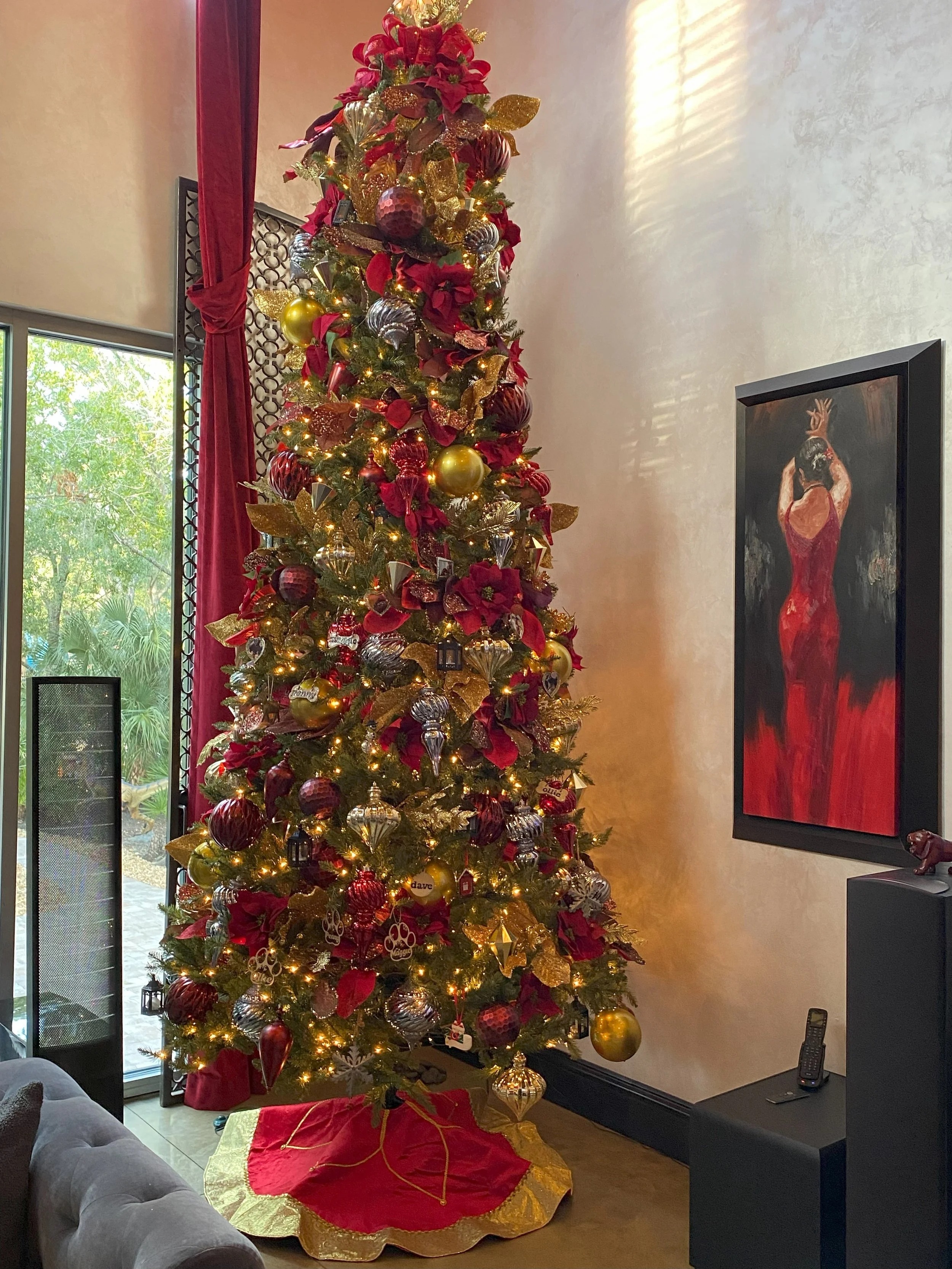 Beautiful decorated Christmas tree with gold, red, and silver ornaments, poinsettia flowers, and lights, in a living room near a window with red curtains.
