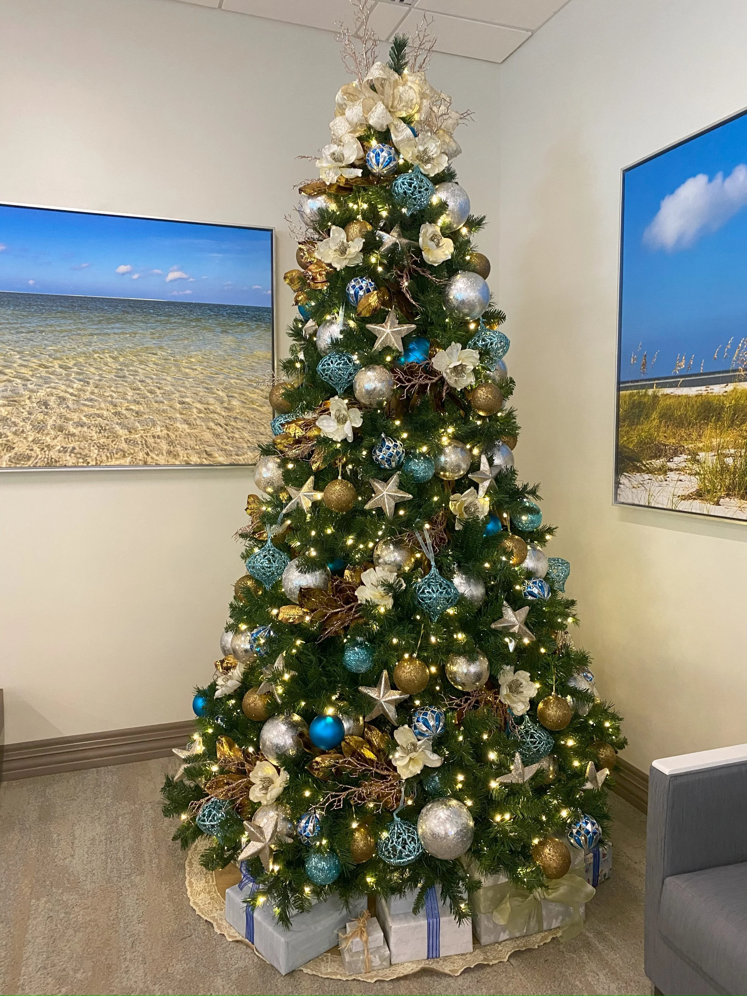 Decorated Christmas tree with gold, silver, and blue ornaments, white flowers, and string lights, with wrapped presents underneath.