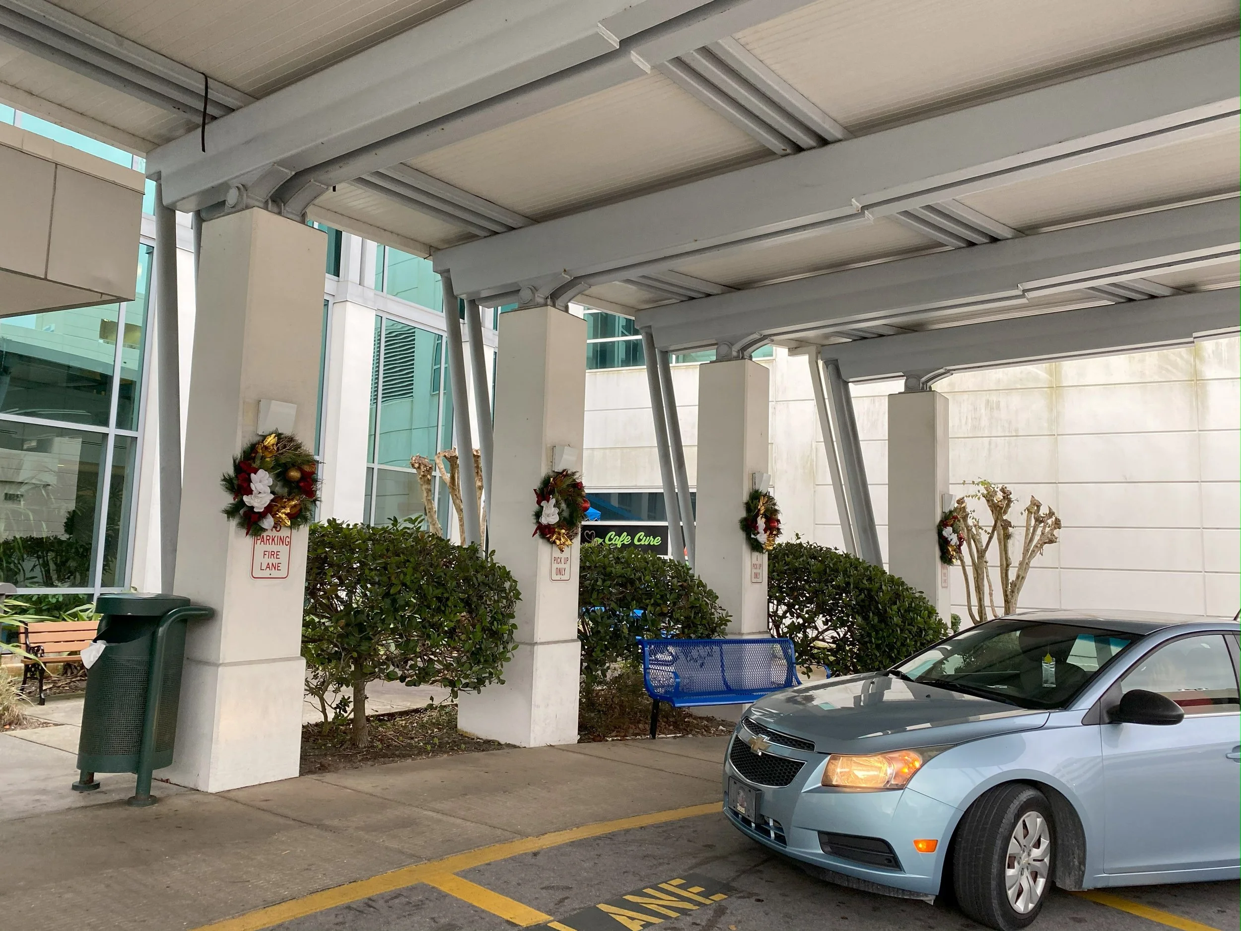 Parking area with a silver car, decorated Christmas wreaths on the pillars, blue bench, and a trash can under a covered walkway outside a modern building.