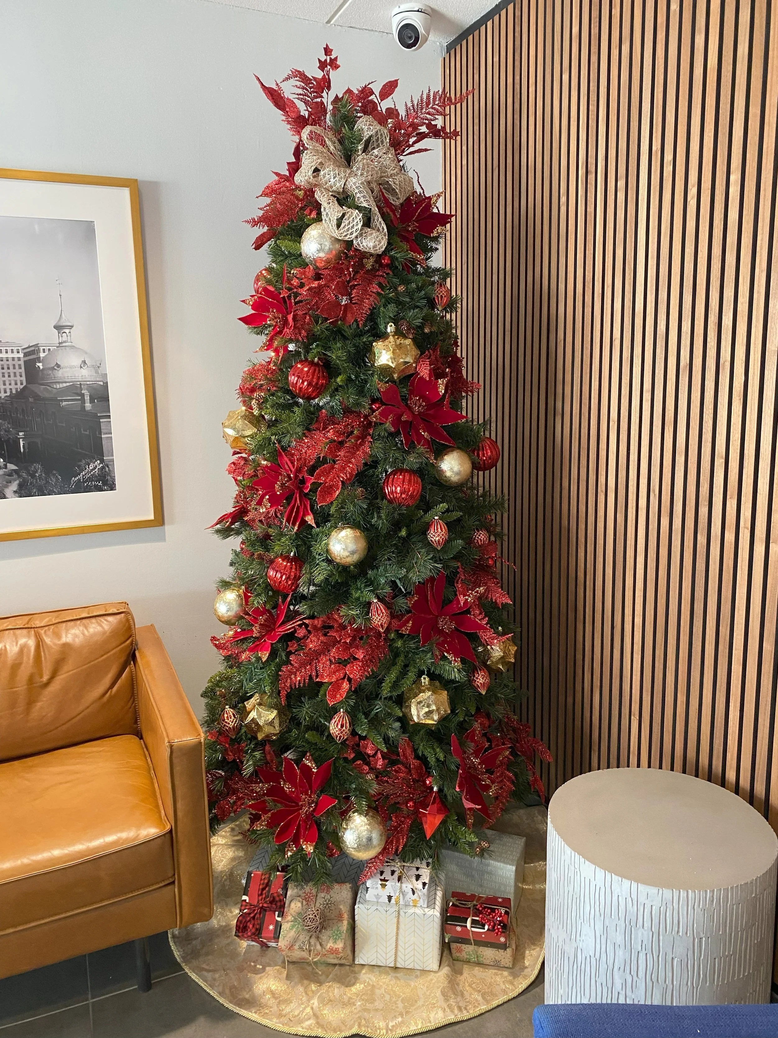 Decorated Christmas tree with red, gold, and glittery ornaments, poinsettia flowers, wrapped presents underneath, adjacent to a leather sofa and a wooden-paneled wall.