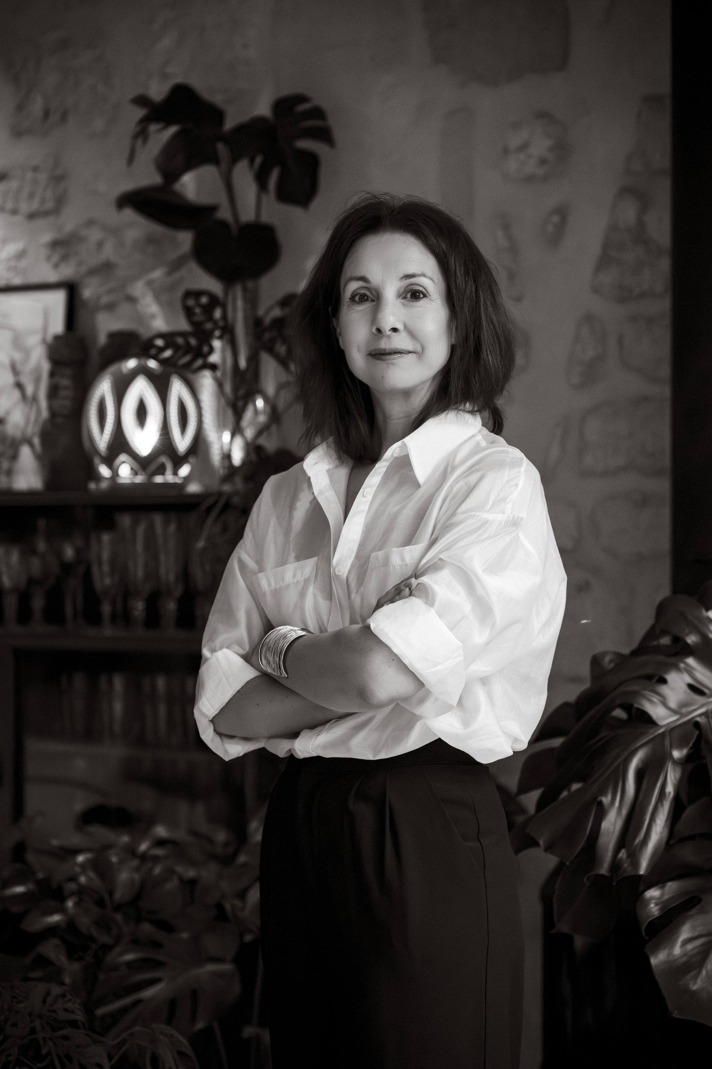 Portrait en noir et blanc d'une femme aux cheveux courts et cheveux bouclés, portant une chemise blanche à manches roulées, croisée sur sa poitrine, dans un intérieur décoré avec des plantes et des objets artistiques.