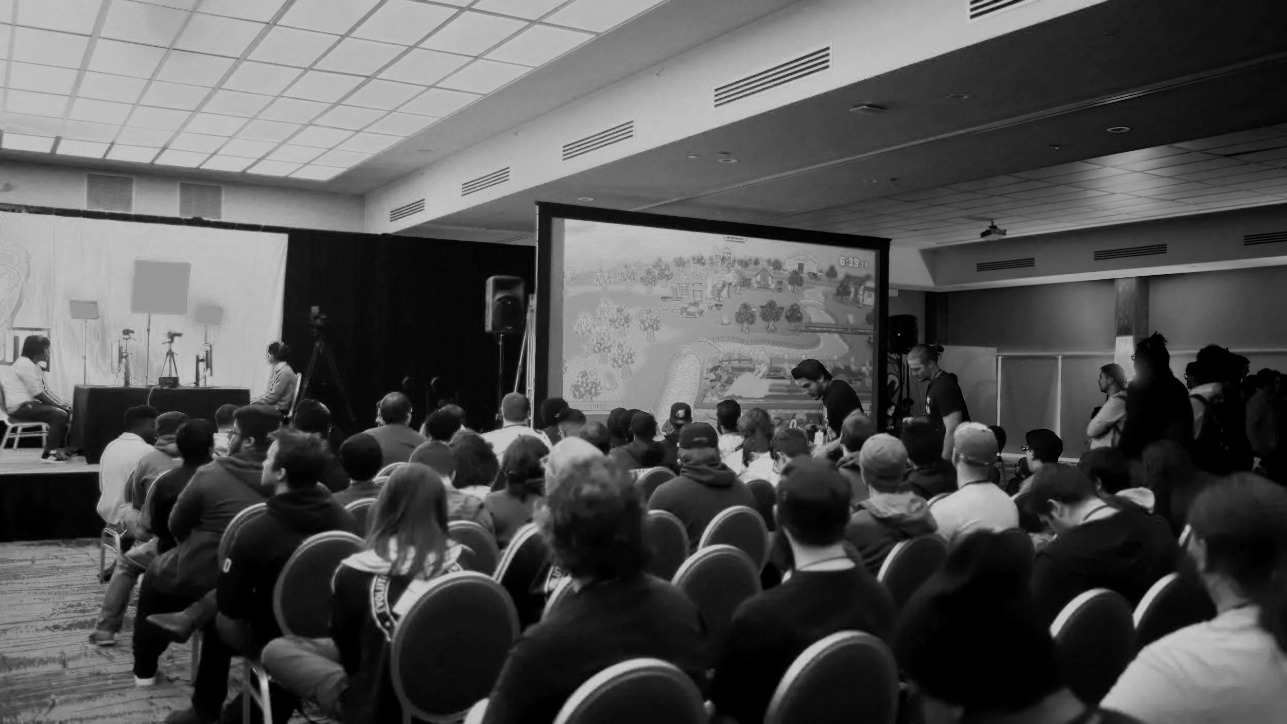 A large group of people seated facing a stage in a conference room with blue lighting, watching a video game being played on a large screen.