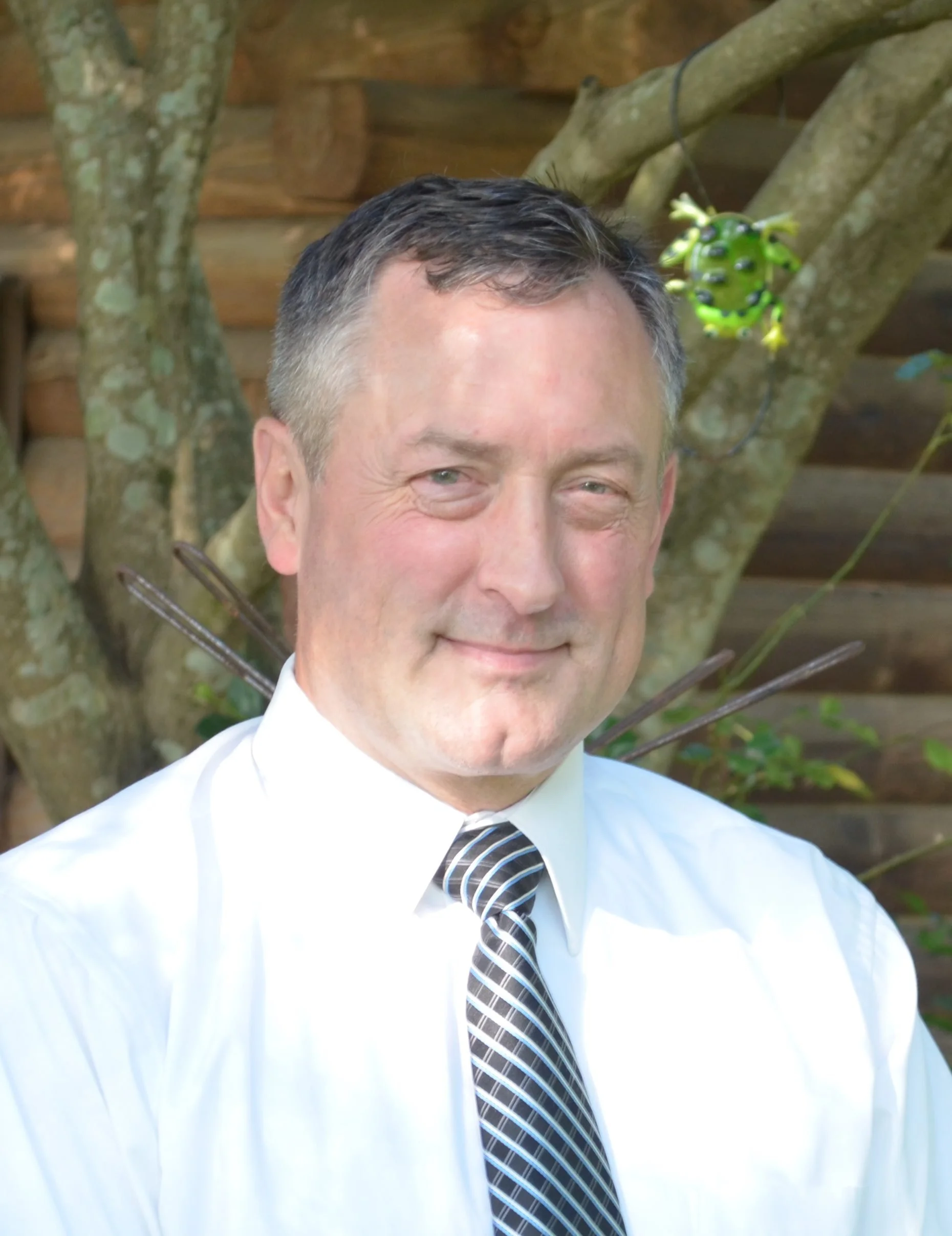 A man with short dark hair, wearing a white shirt and a striped tie, smiling outdoors with trees and a wooden fence in the background.