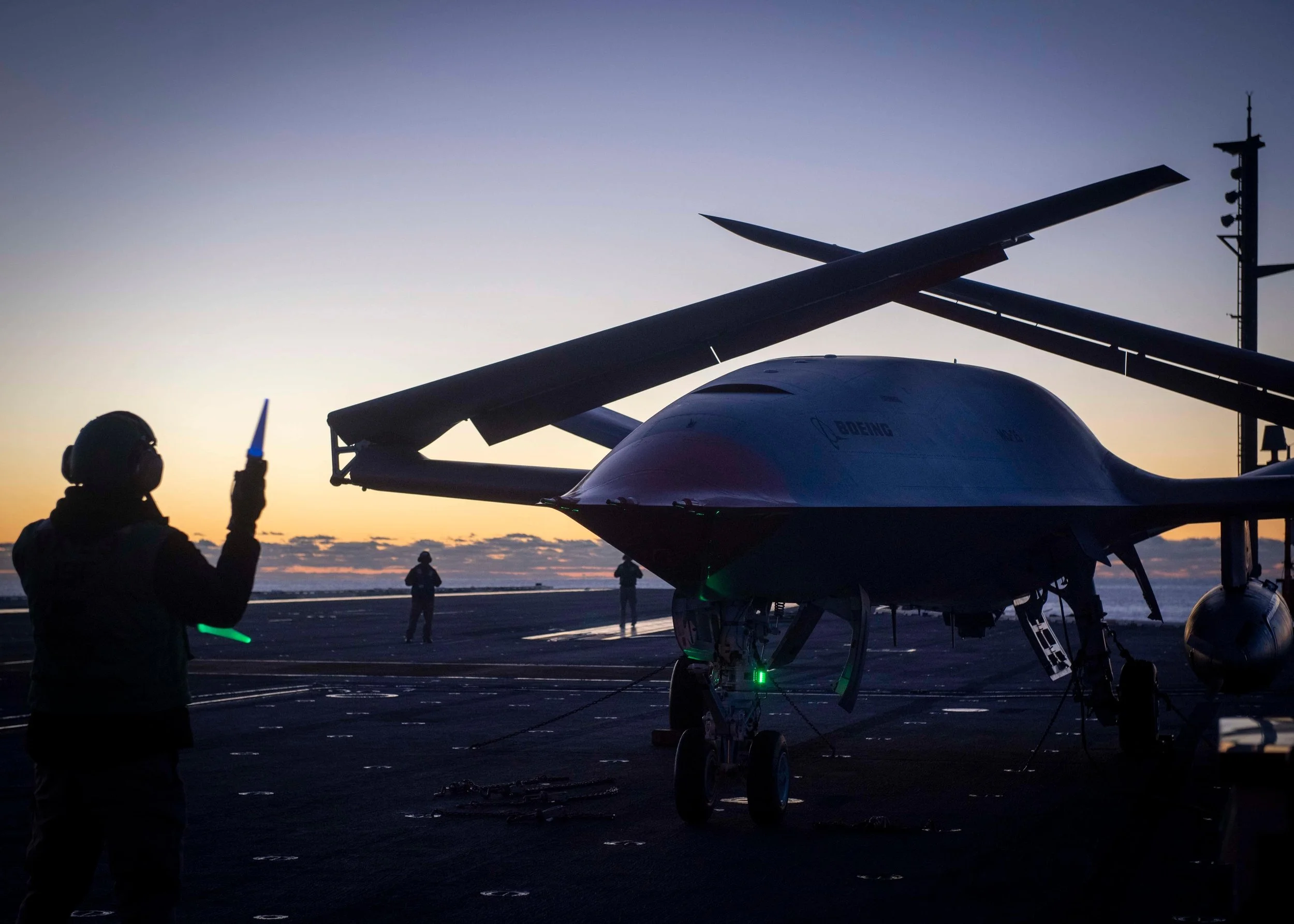 Silhouette of a person holding a light stick on an aircraft carrier at sunset, with a drone aircraft and crew members in the background.
