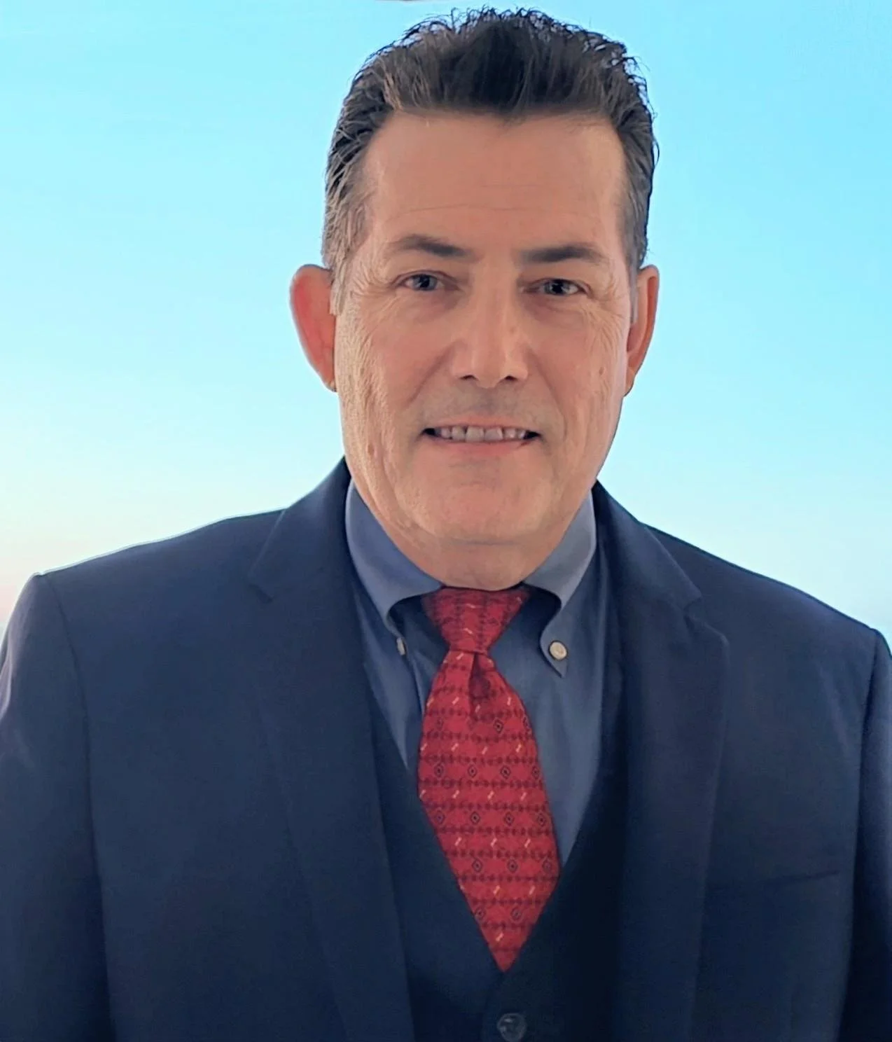 A man wearing a dark suit, blue shirt, and red patterned tie, smiling slightly against a clear blue sky background.