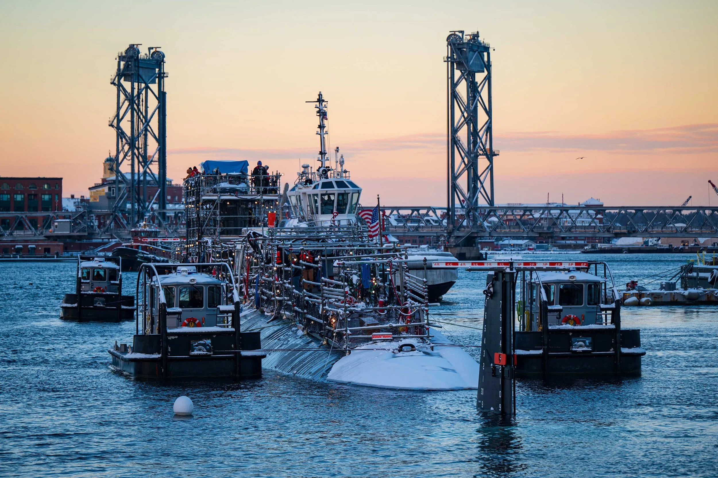 Submarine docked in harbor with a bridge in the background during sunset.
