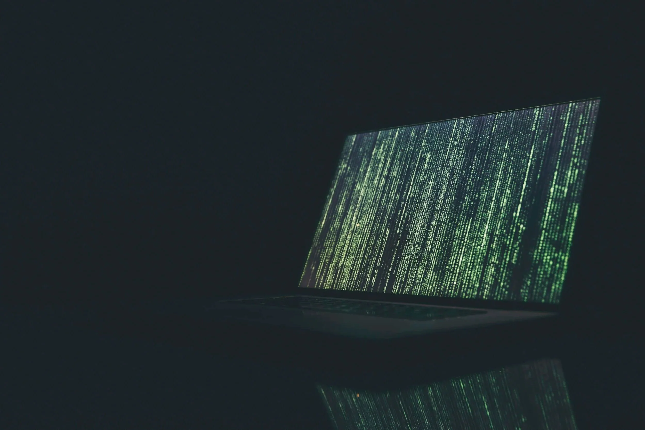 A laptop computer with the screen displaying green digital code on a dark background, reflecting on a glossy surface.