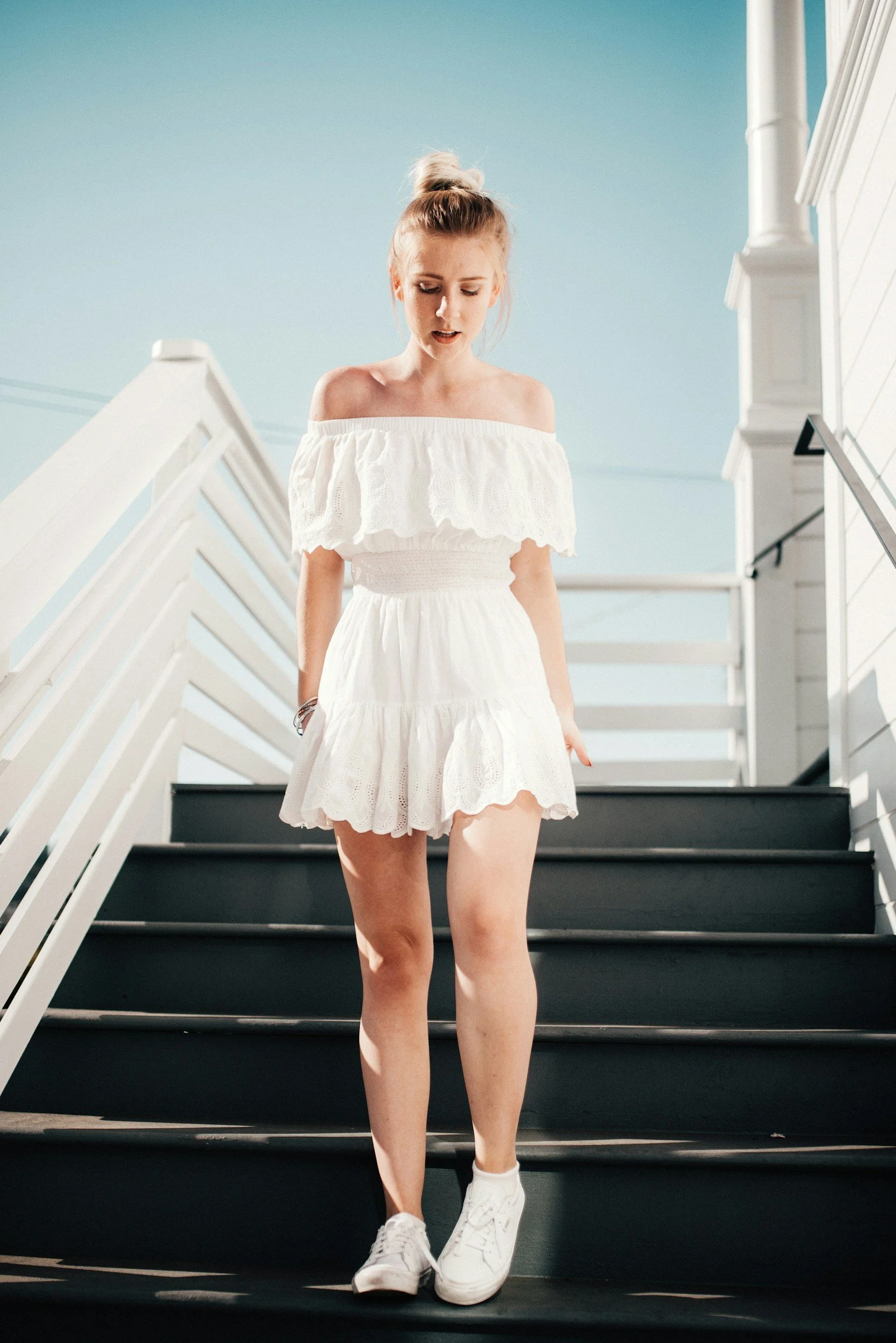 Woman in a white off-the-shoulder dress walking down outdoor stairs in bright sunlight.