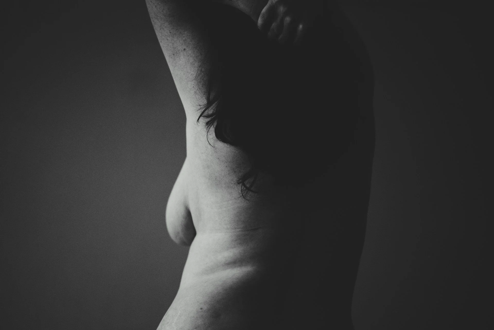 Black-and-white rear side profile of a shirtless woman with one arm raised.