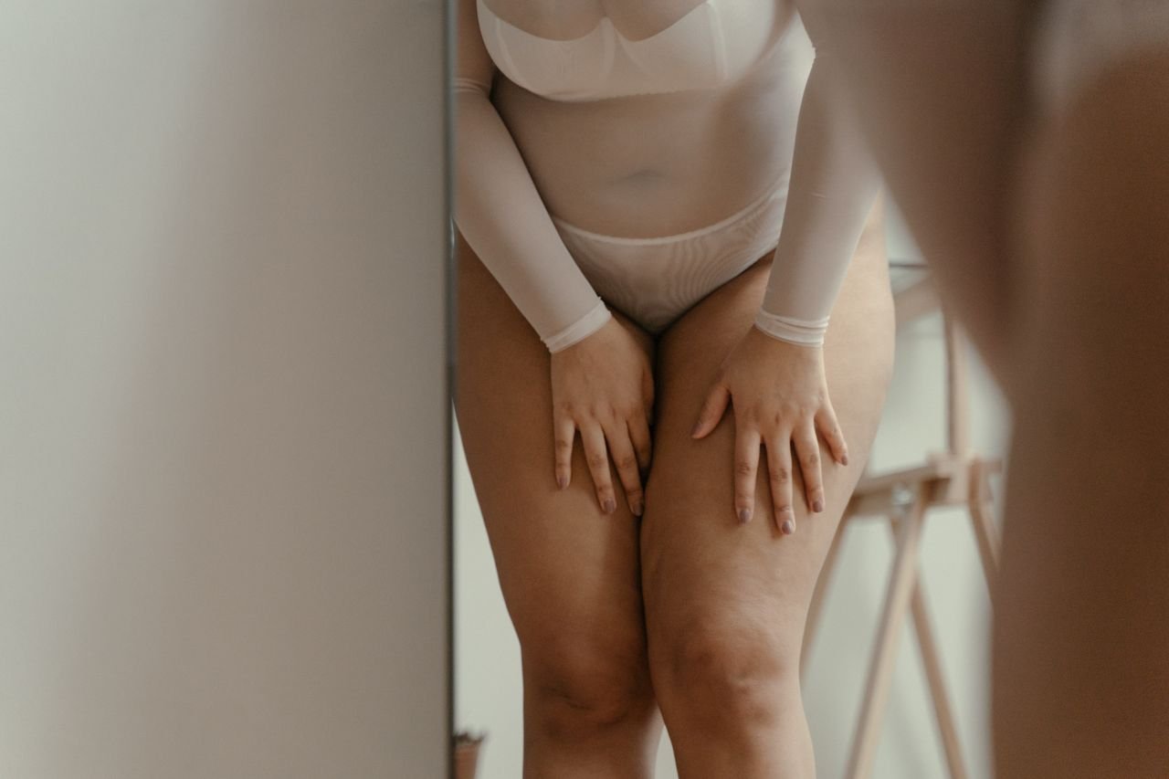 Woman in a light-colored onesie underwear looking at their body in a full-length mirror.