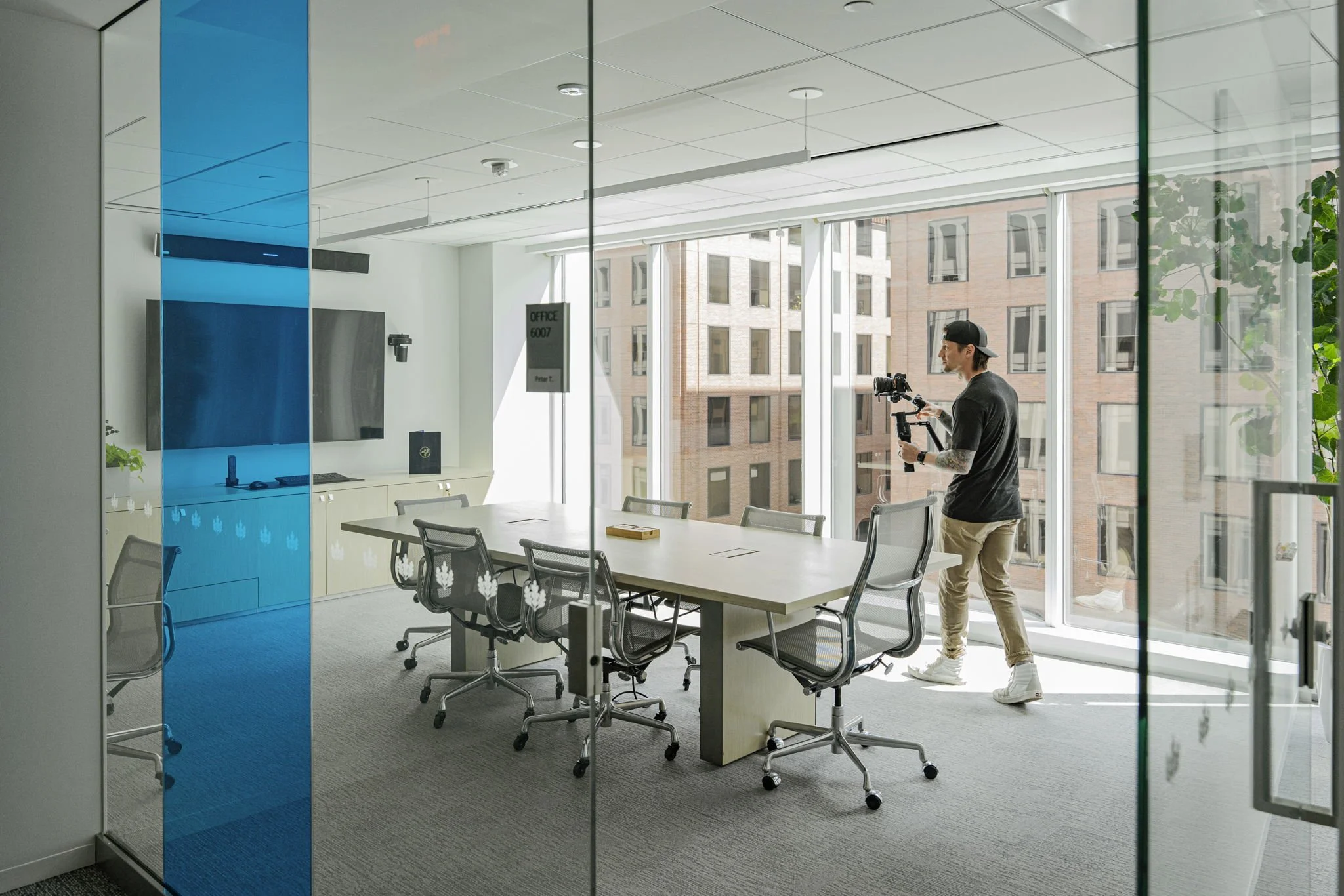 A man setting up video equipment in a modern conference room with glass walls, a long table, and office chairs, with large windows showing a cityscape.