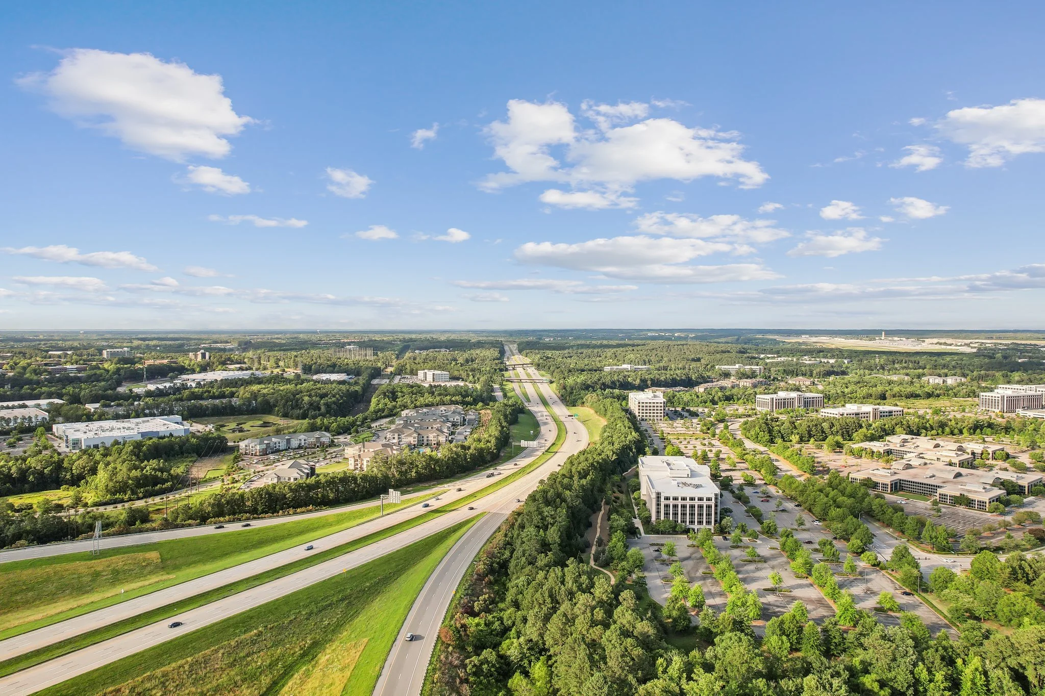 An aerial view of a highway cutting through a green landscape with buildings and trees, under a blue sky with scattered clouds.