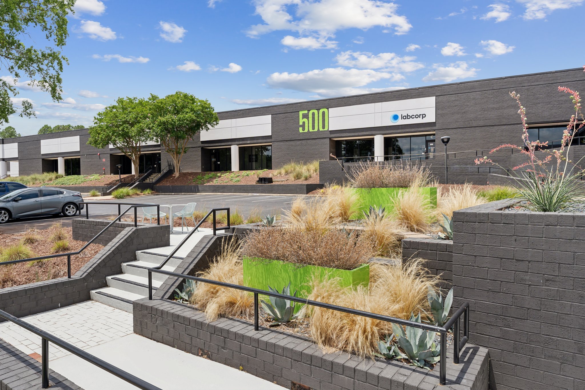 Modern office building with a parking lot and landscaped garden in the front, featuring drought-tolerant plants and trees.