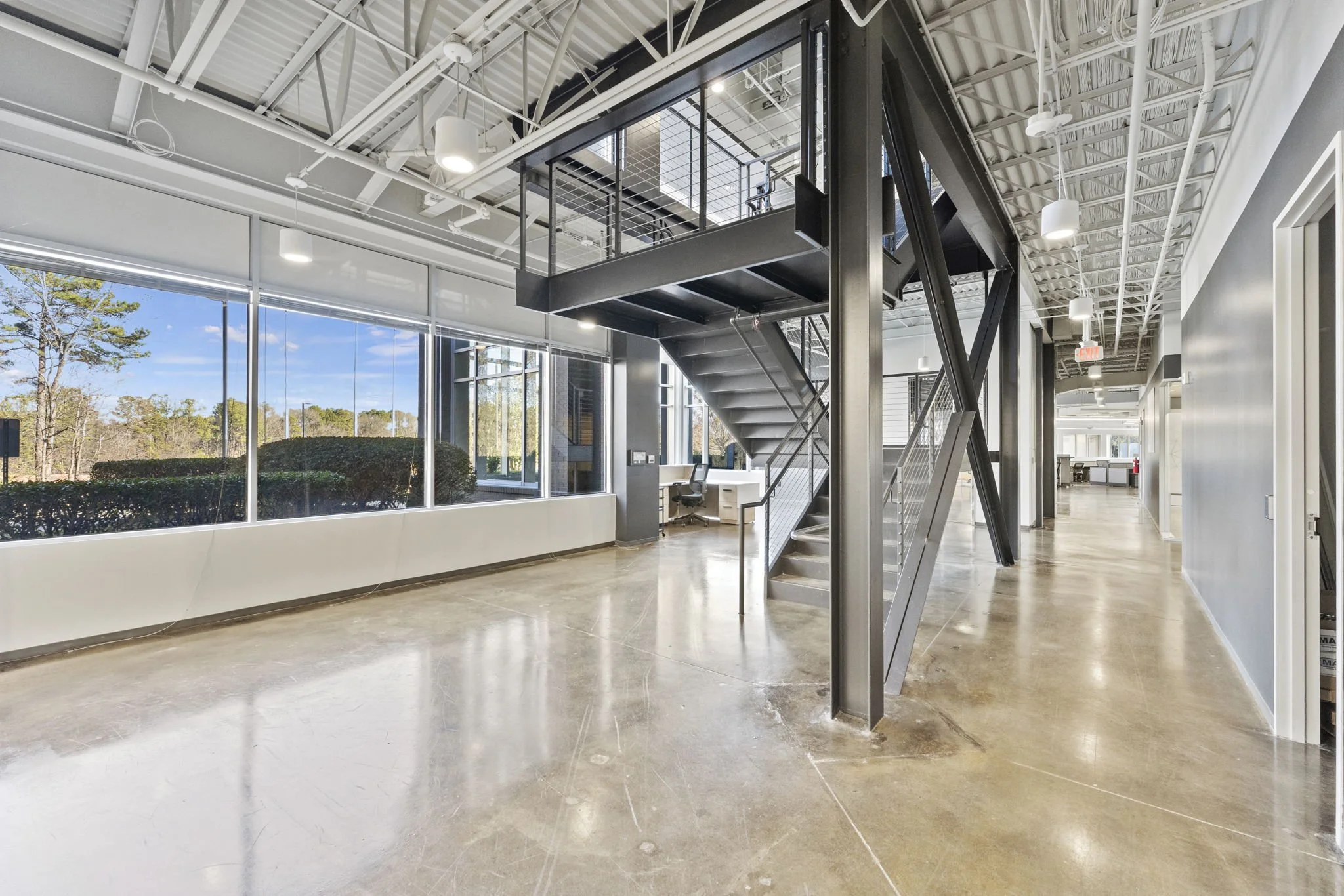 Modern office space with large windows, polished concrete floors, and a metal staircase leading to a mezzanine level.