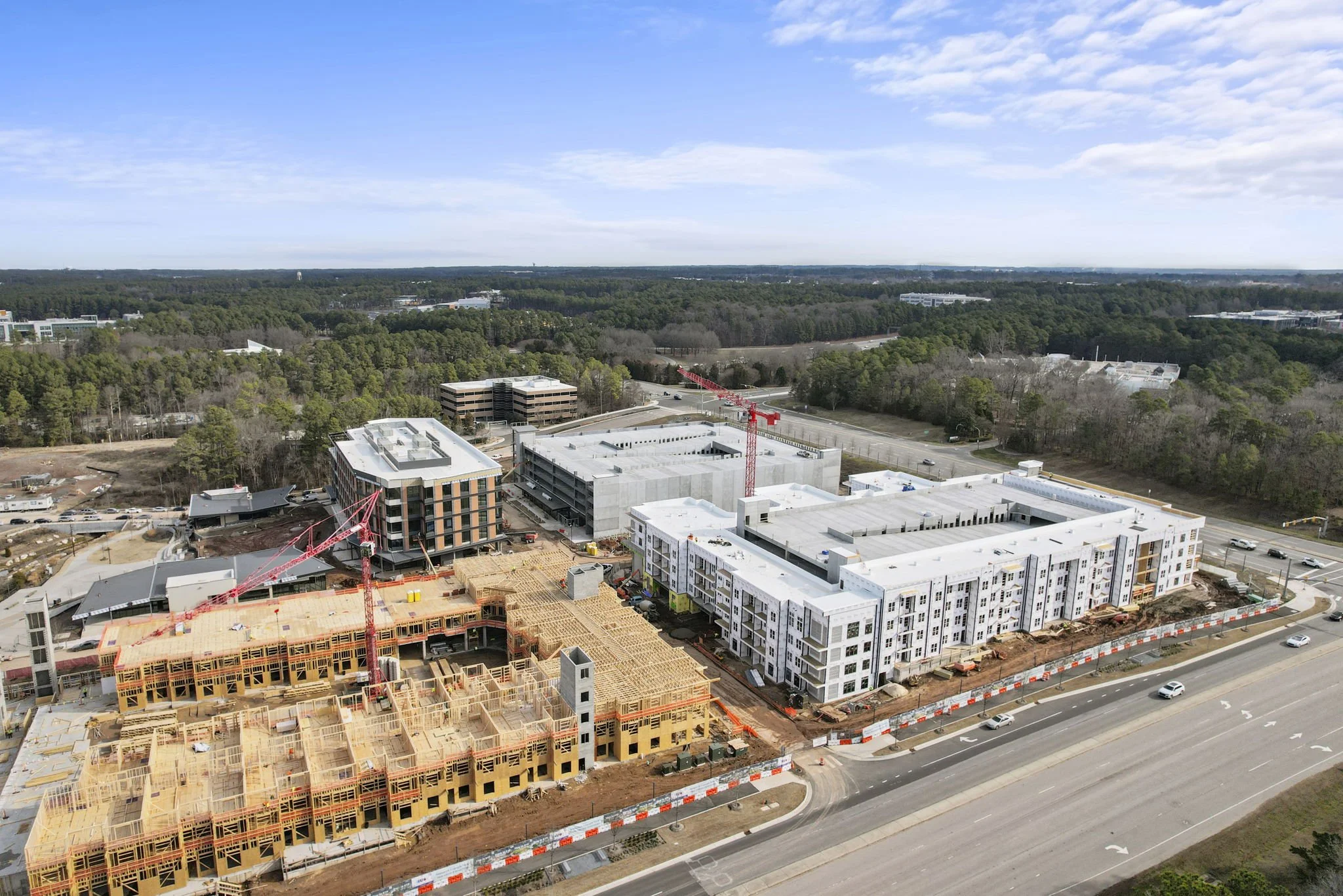 Aerial view of a large building construction site with multiple cranes and partially built structures, surrounded by roads and wooded areas in the background.