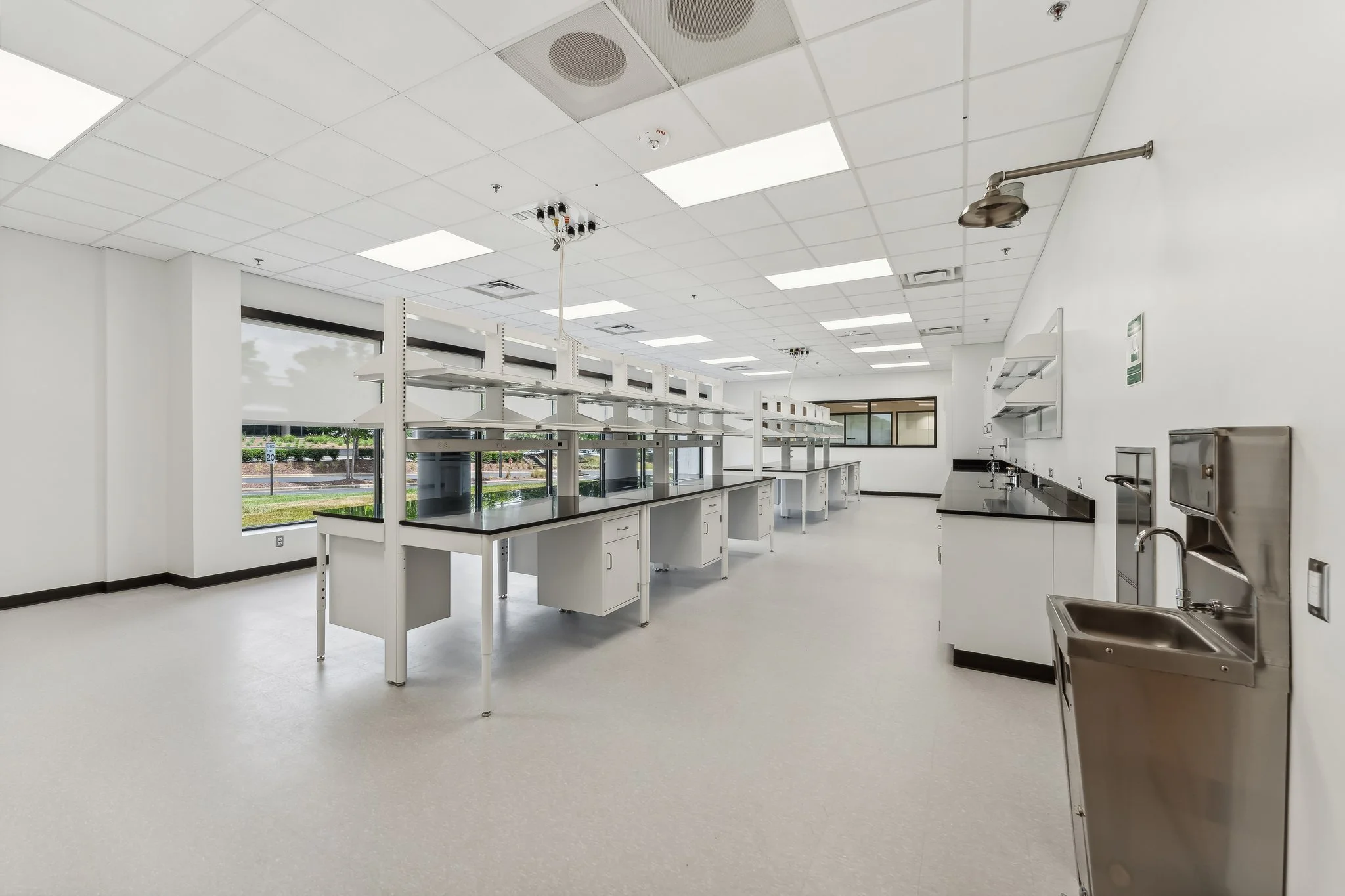 Empty commercial kitchen with white walls, black countertops, stainless steel sink, and storage shelves, illuminated by ceiling lights with windows showing outside view.