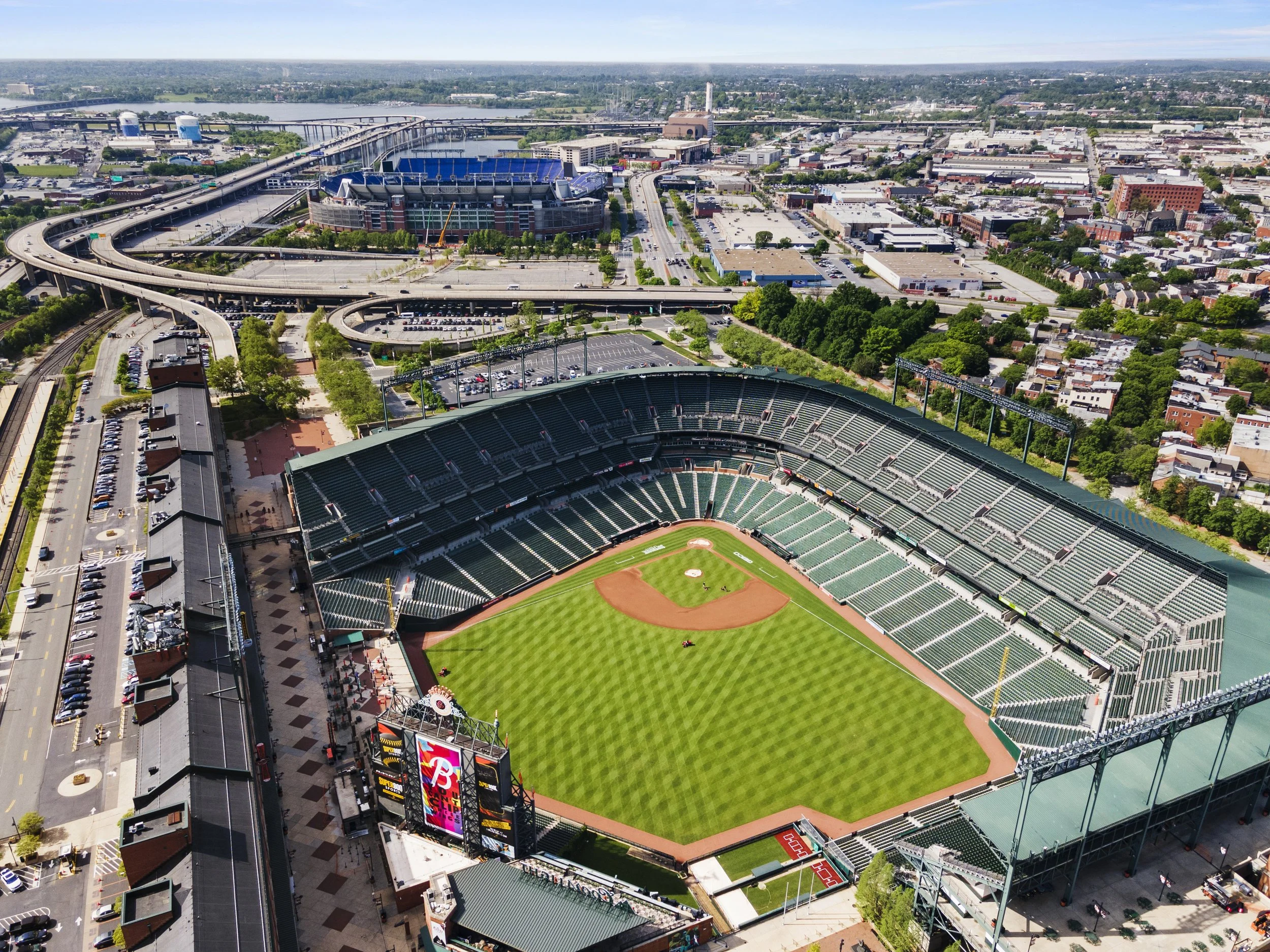 Aerial view of a baseball stadium in a city, with green field, seating areas, adjacent parking lots, highways, and urban landscape in the background.