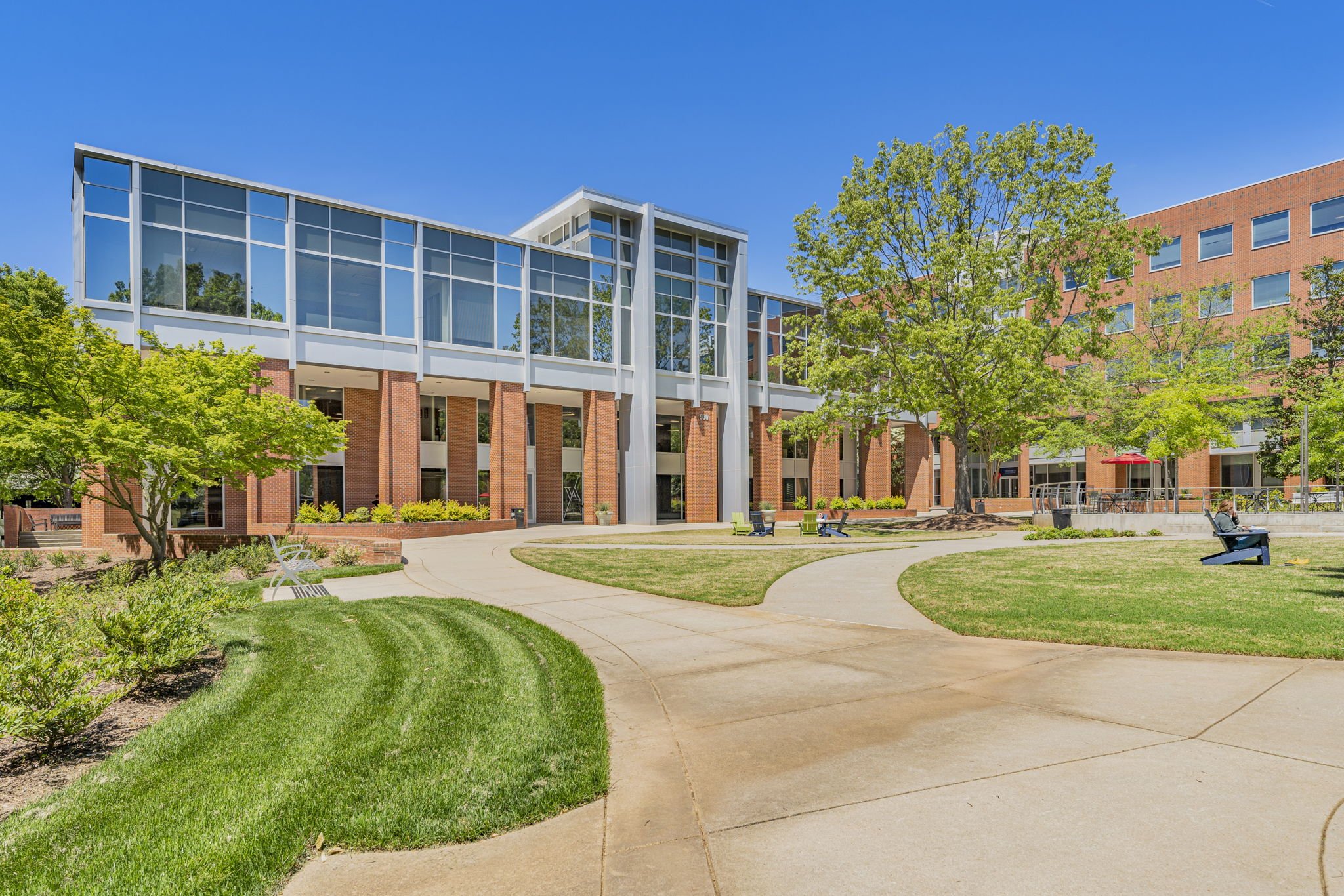 A modern multi-story building with large glass windows, surrounded by a landscaped courtyard with green grass, trees, benches, and a clear blue sky.
