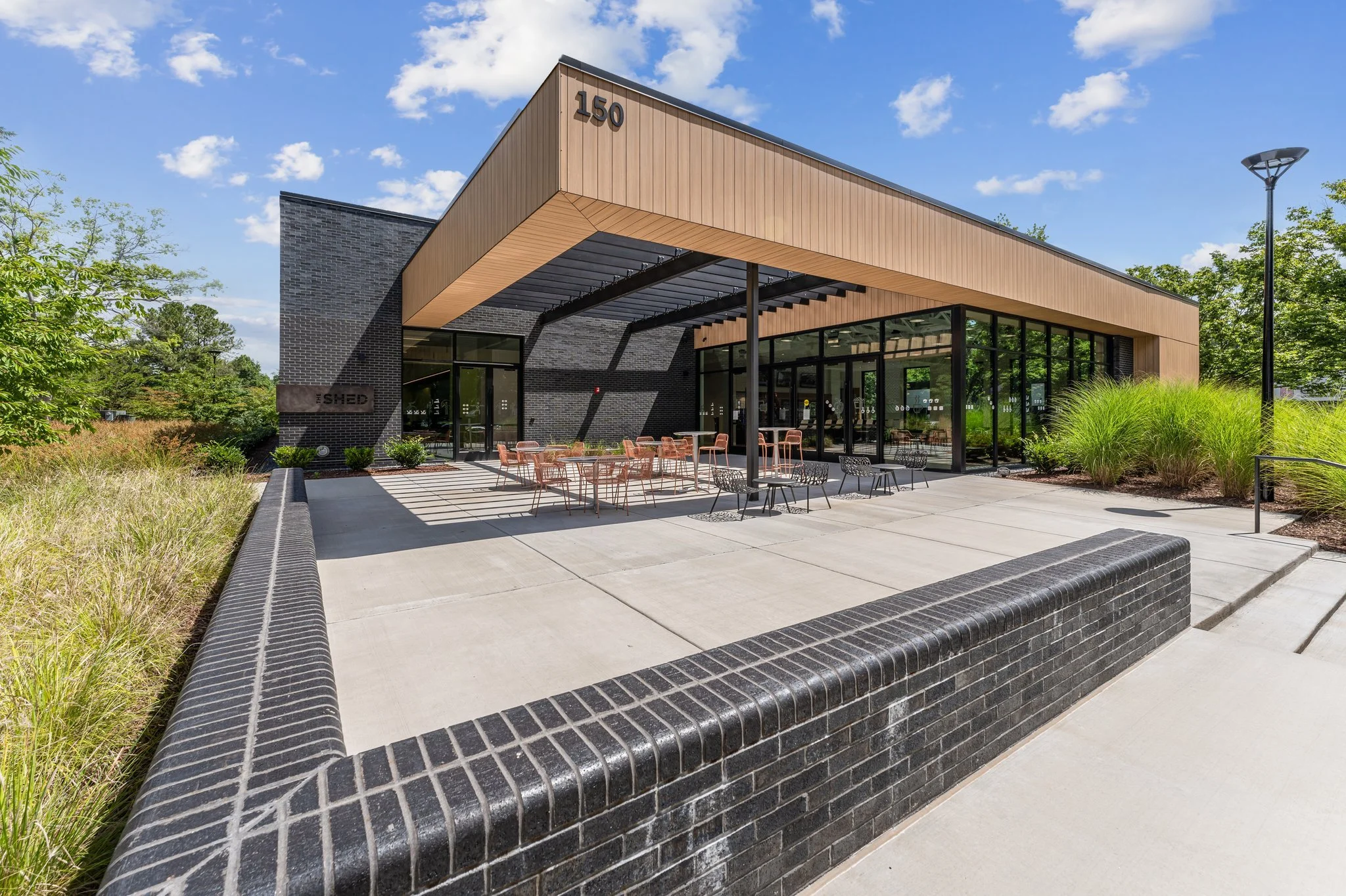 Modern building with a large covered outdoor seating area, glass walls, and surrounding landscaping on a sunny day.