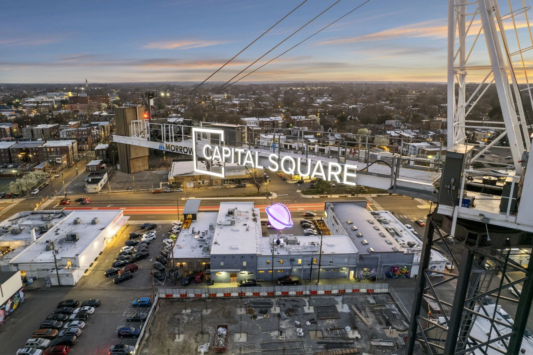 A nighttime cityscape view from a rooftop with a large illuminated sign reading "CAPITAL SQUARE" and a crane structure on the right. The city has buildings and streets with moving cars, and the sky shows a colorful sunset.