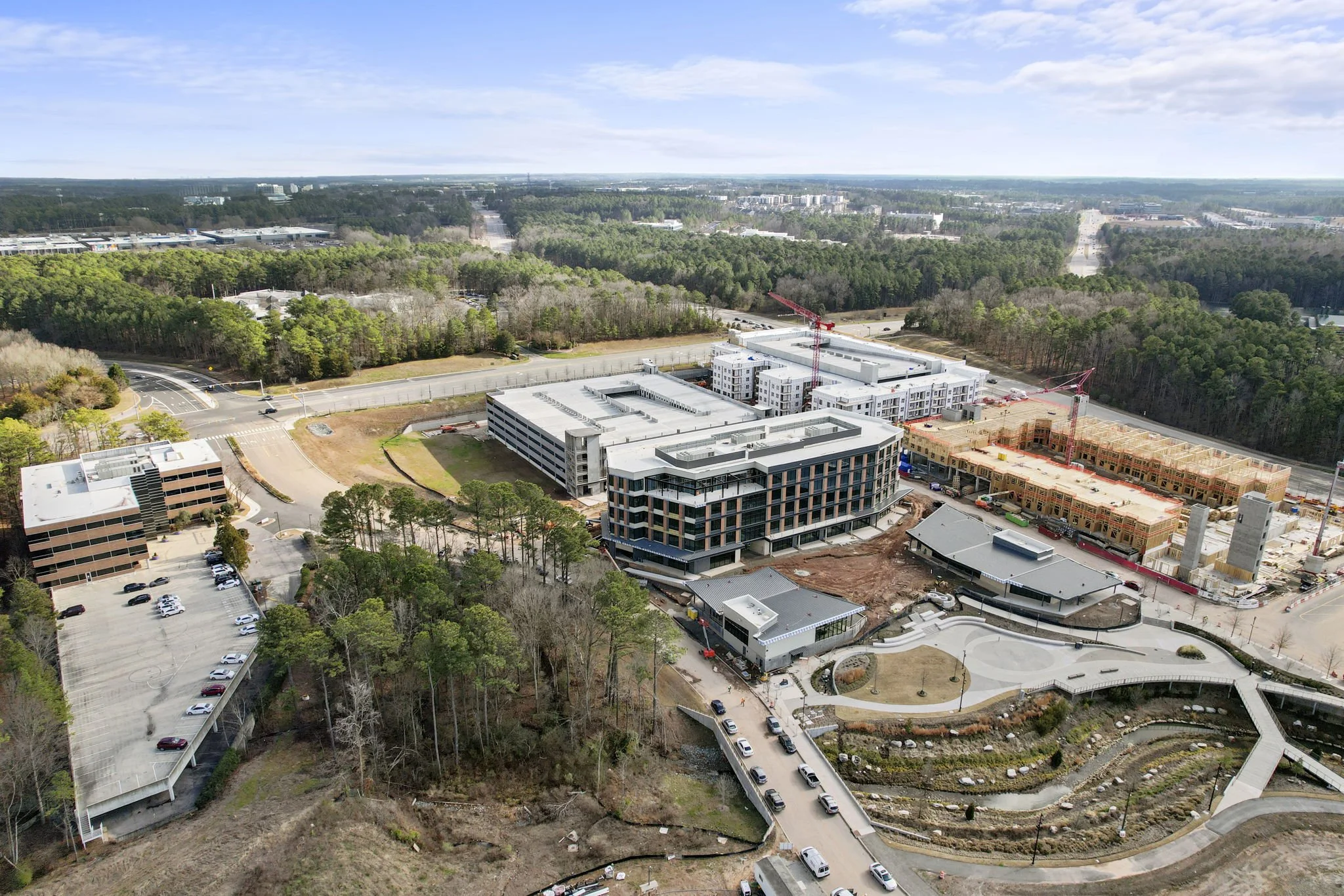 An aerial view of a construction site with several buildings in progress, two cranes, parking lots, and surrounding trees under a partly cloudy sky.