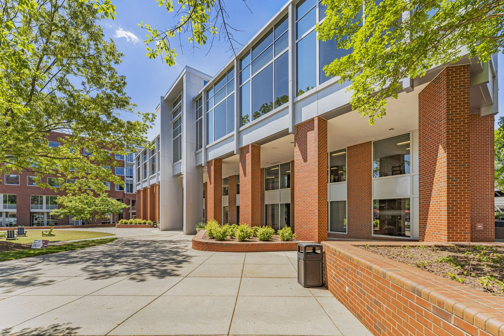 Exterior view of a modern multi-story building with brick and glass facade, surrounded by trees and a courtyard with benches and a trash can.