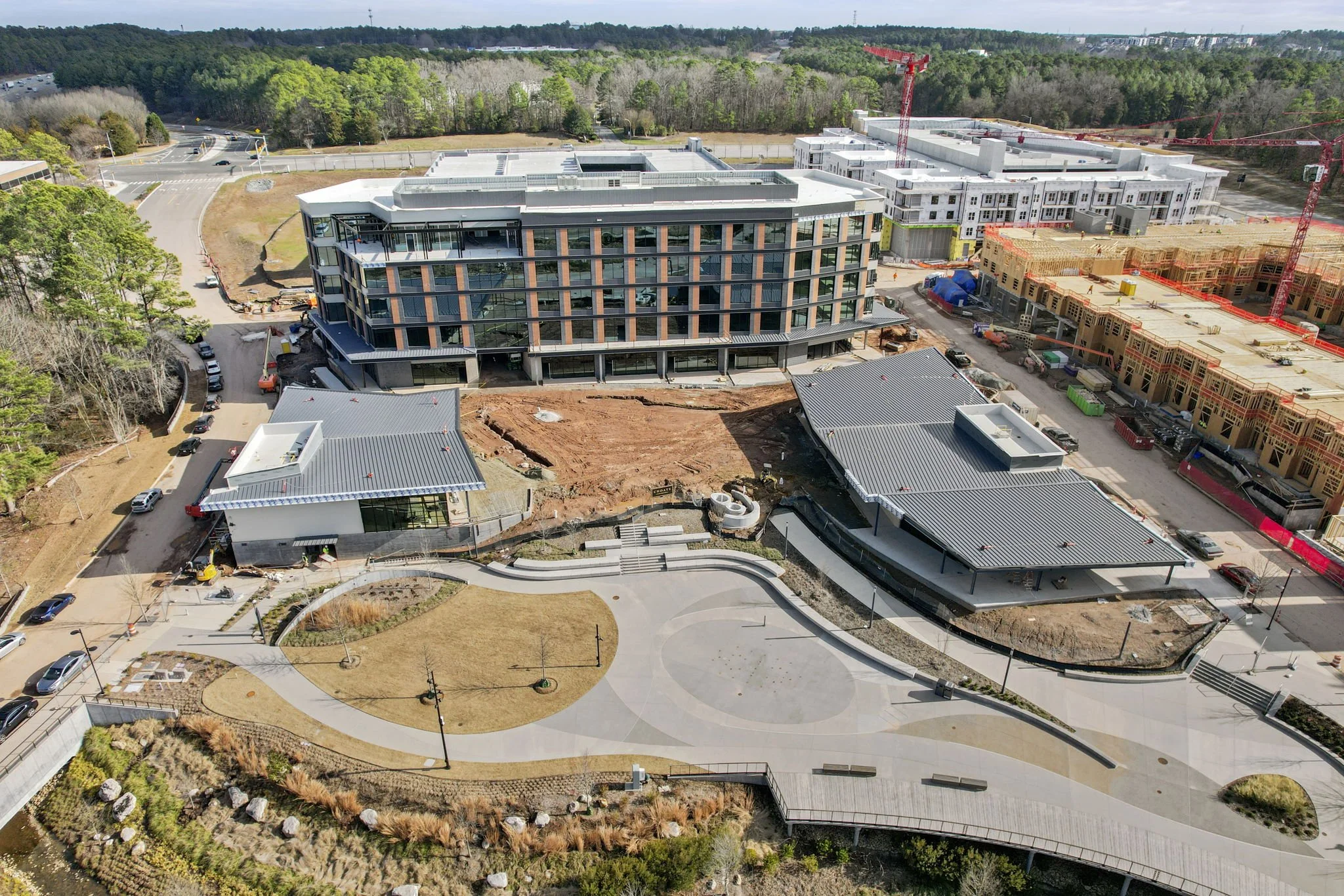 An aerial view of a construction site with multiple buildings in various stages of completion, surrounded by trees and roads.