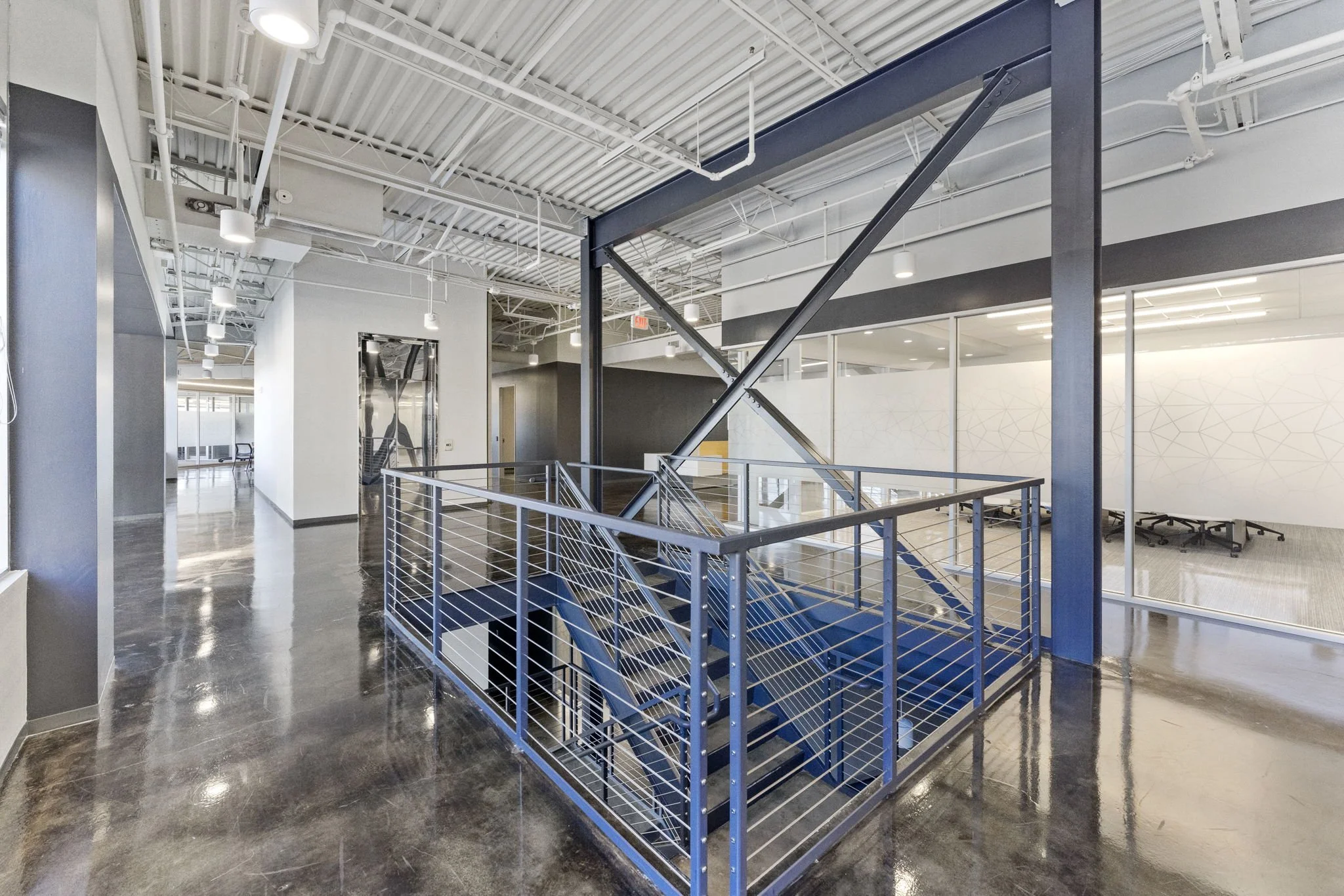 Modern office interior with a staircase enclosed by a metal railing, glass walls, and exposed ceiling pipes.