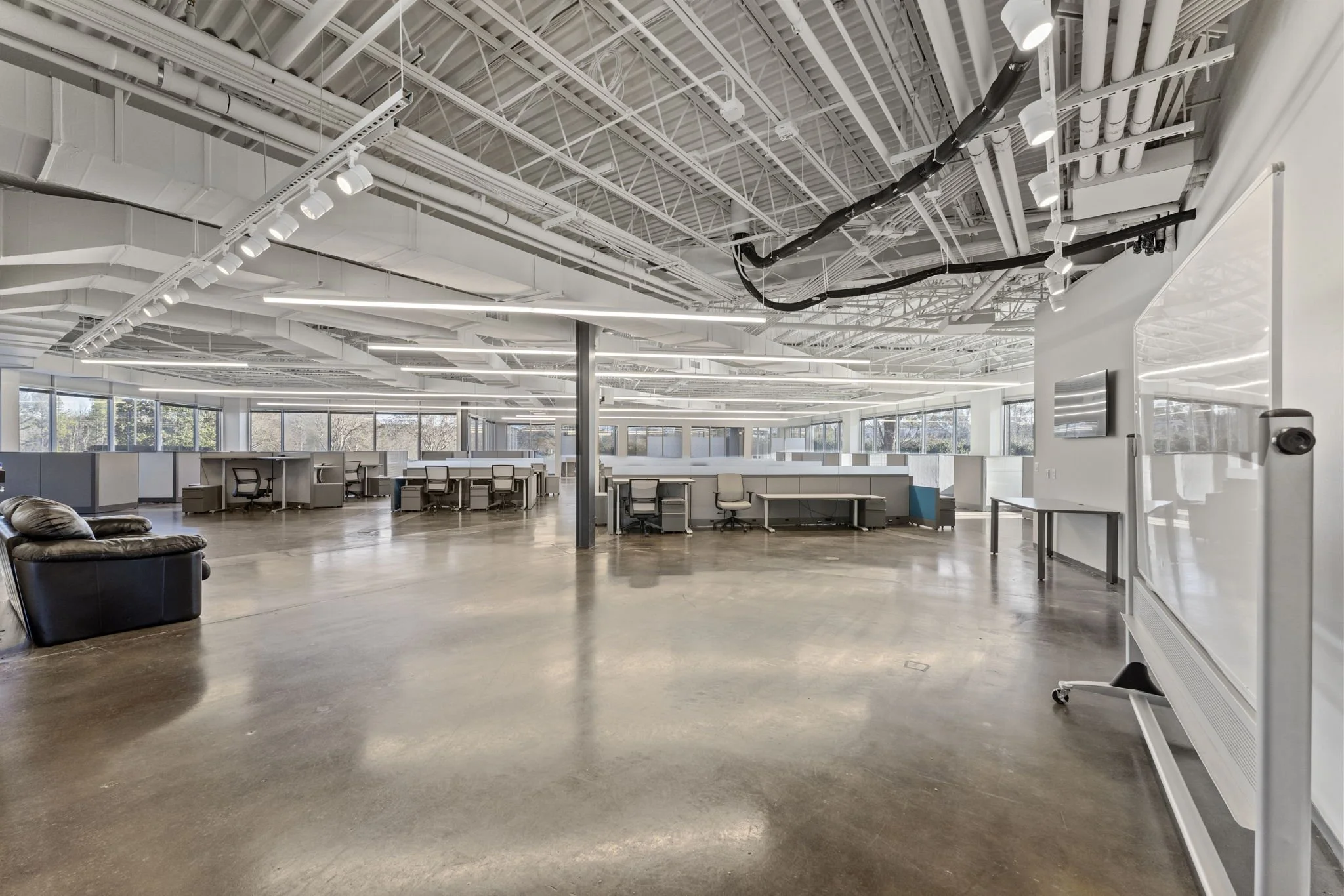Empty modern office space with large windows, desks, chairs, a whiteboard, and a sofa.