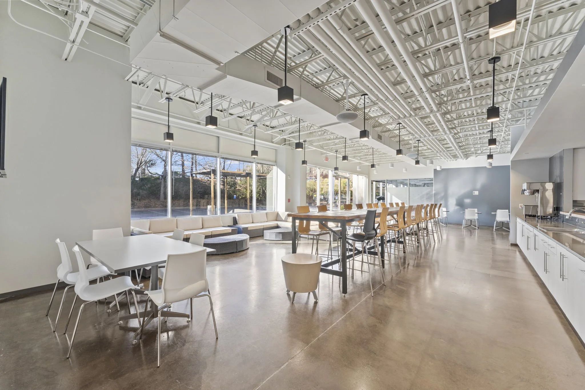 Interior of a modern lounge or cafeteria with large windows, white walls, and a high, exposed ceiling with black pendant lights. There are white tables with white chairs and a long bar-height table with multiple chairs. A seating area with a sectiona