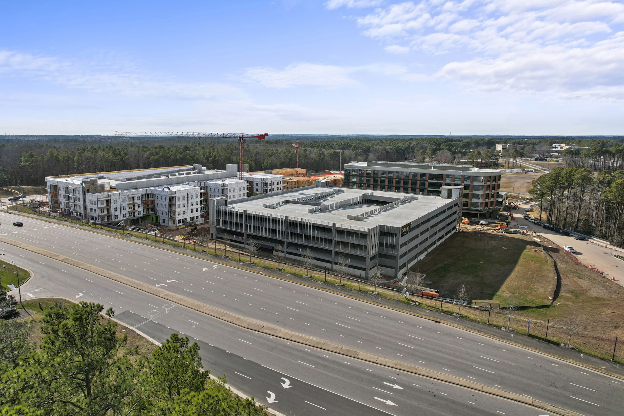 Aerial view of a construction site with multiple buildings, including a parking garage, near a highway, with trees and an open sky in the background.
