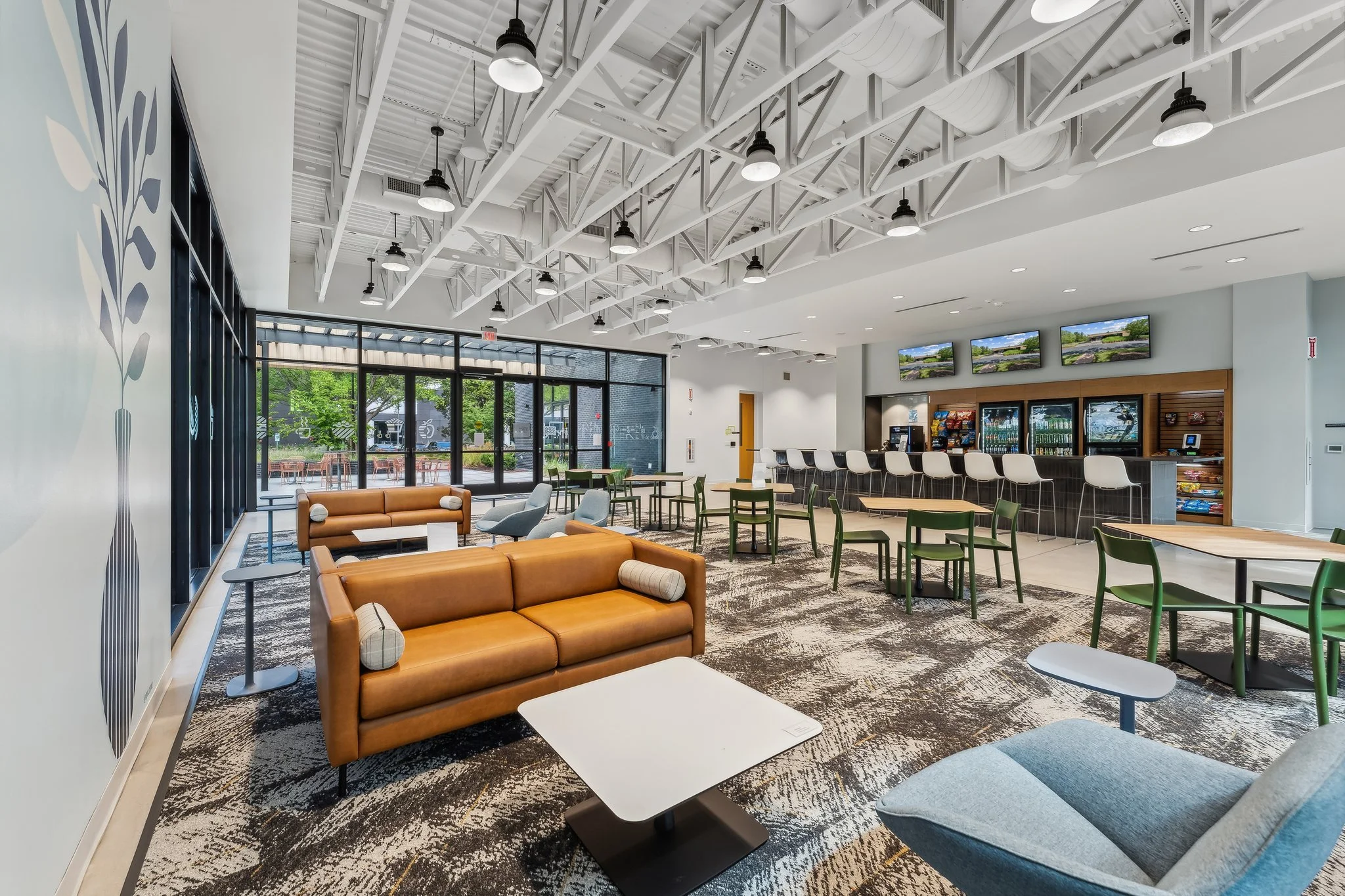 Modern lobby area with brown leather sofas, blue and gray chairs, green and wooden tables, a coffee station, and wall-mounted screens.