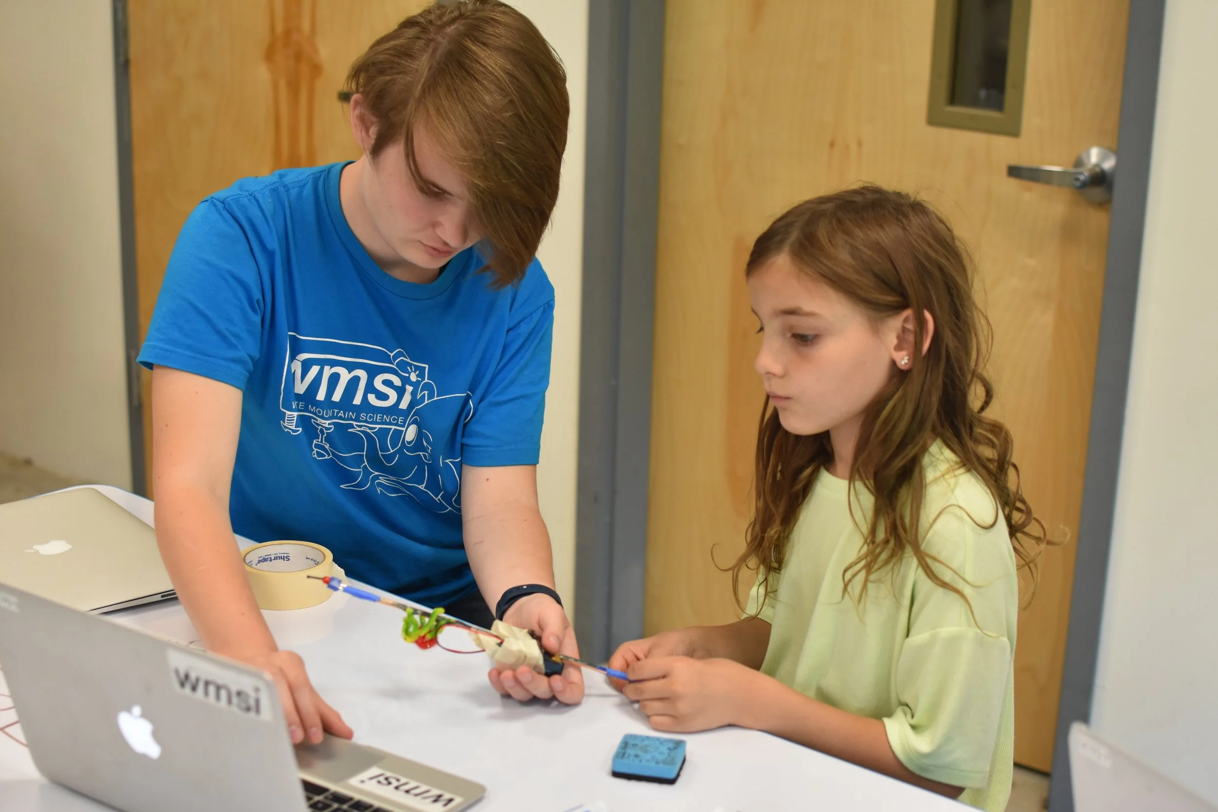 Two girls working together on a science experiment at a table, with a laptop, tape, and a small object in their hands. One girl is wearing a blue t-shirt and the other a yellow shirt.