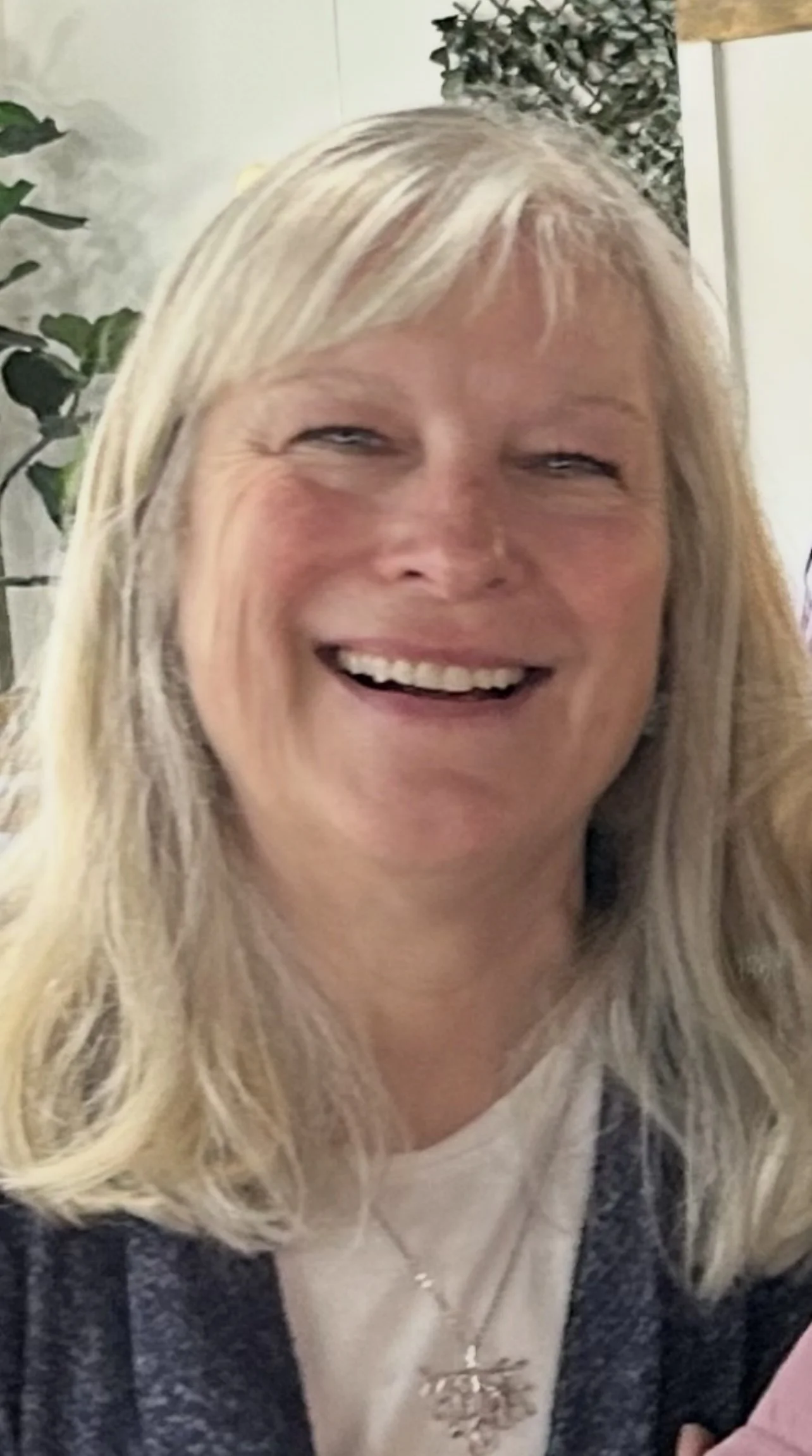 Close-up of a smiling woman with blonde wavy hair, wearing a necklace with a pendant, in an indoor setting with plants in the background.