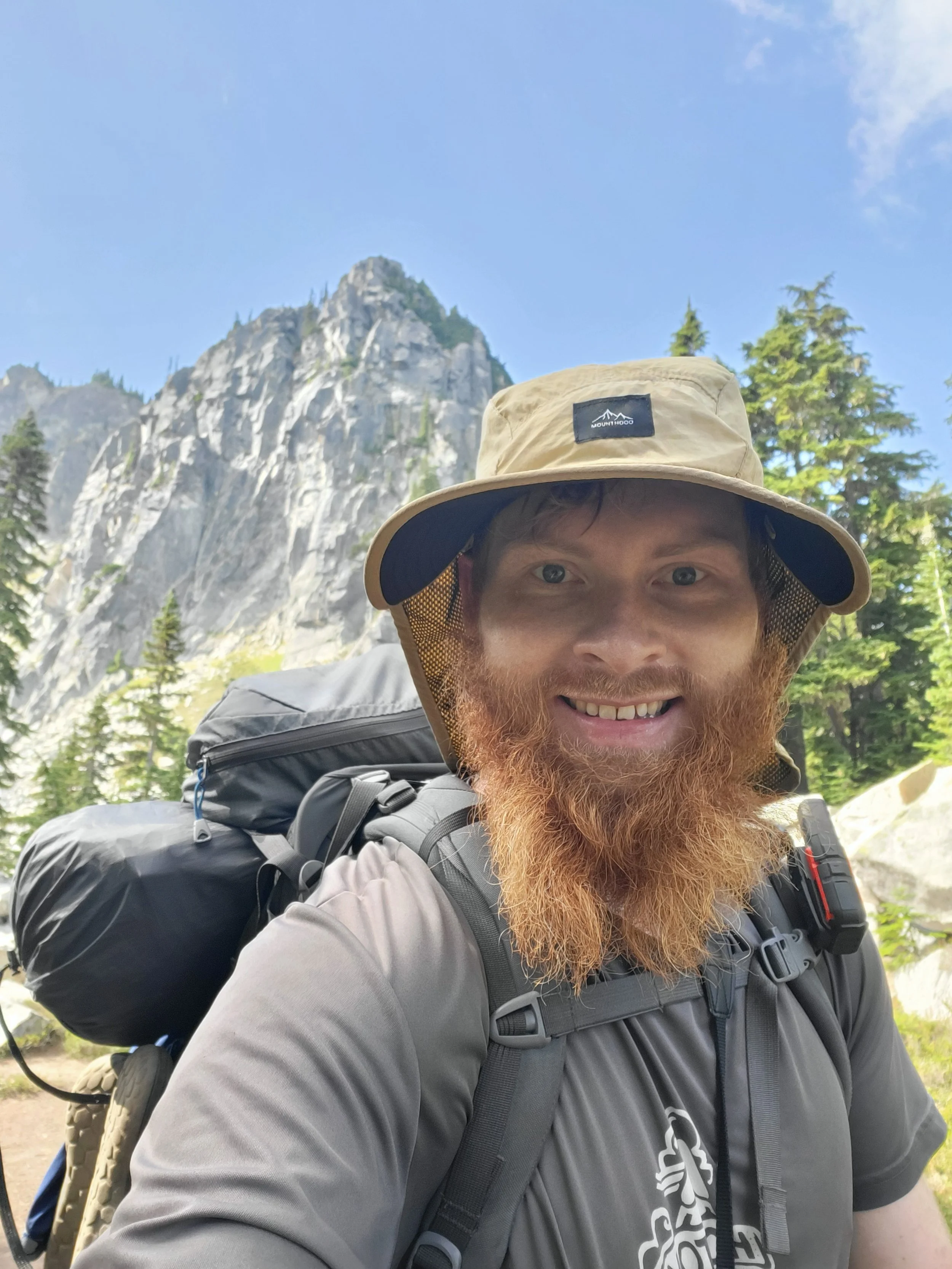A man hiking outdoors, wearing a beige hat and gray outdoor gear, with a large backpack, in a forested area with tall mountains in the background.