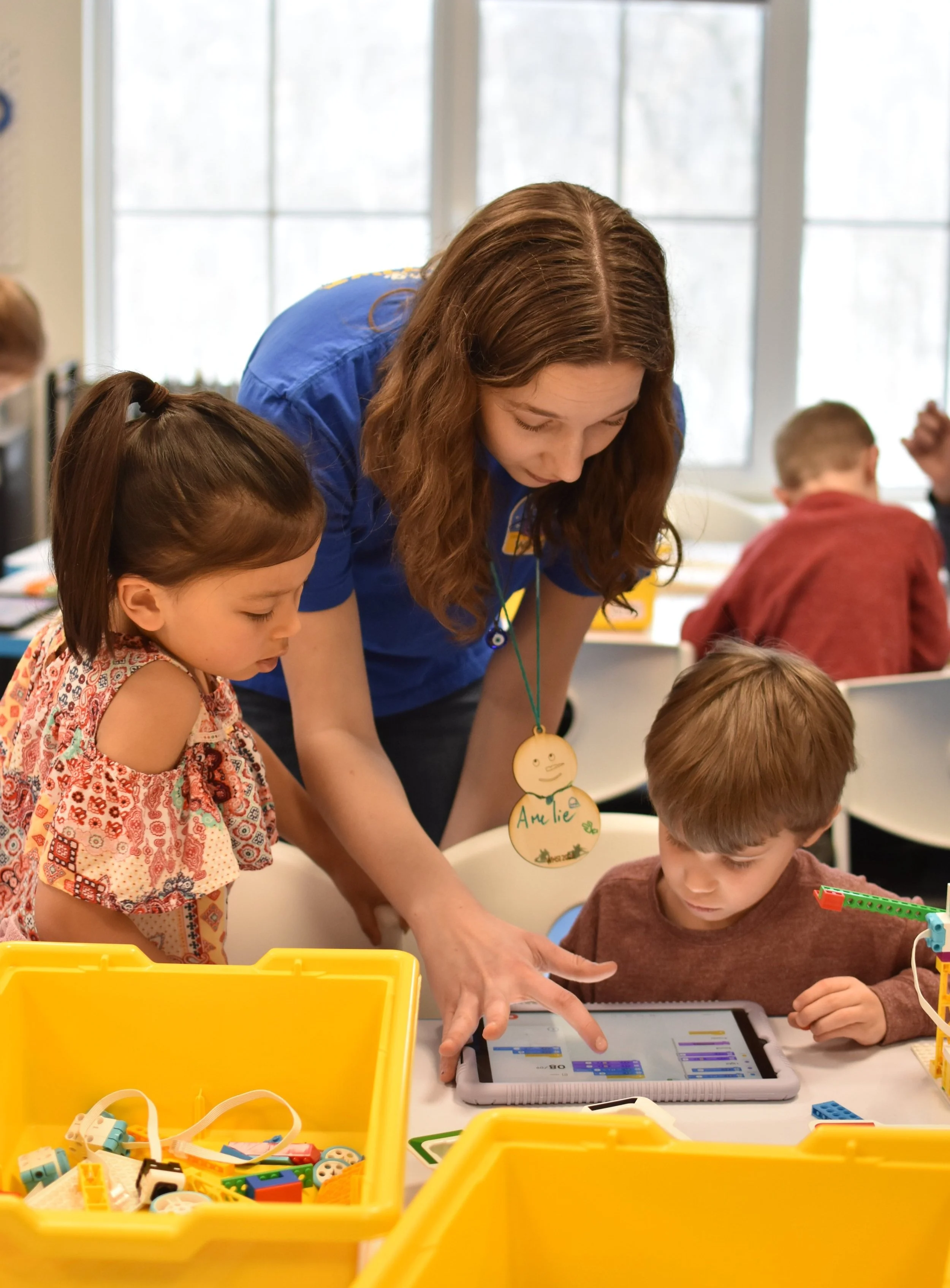 A teacher helps two young students with a tablet computer in a classroom, surrounded by yellow containers of building blocks.