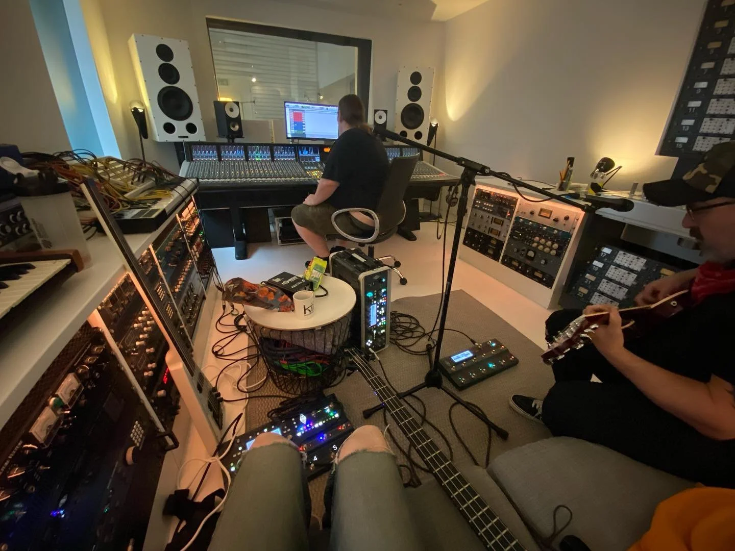 Recording studio setup with mixing console, speakers, guitarists with effects pedals, and audio equipment racks.
