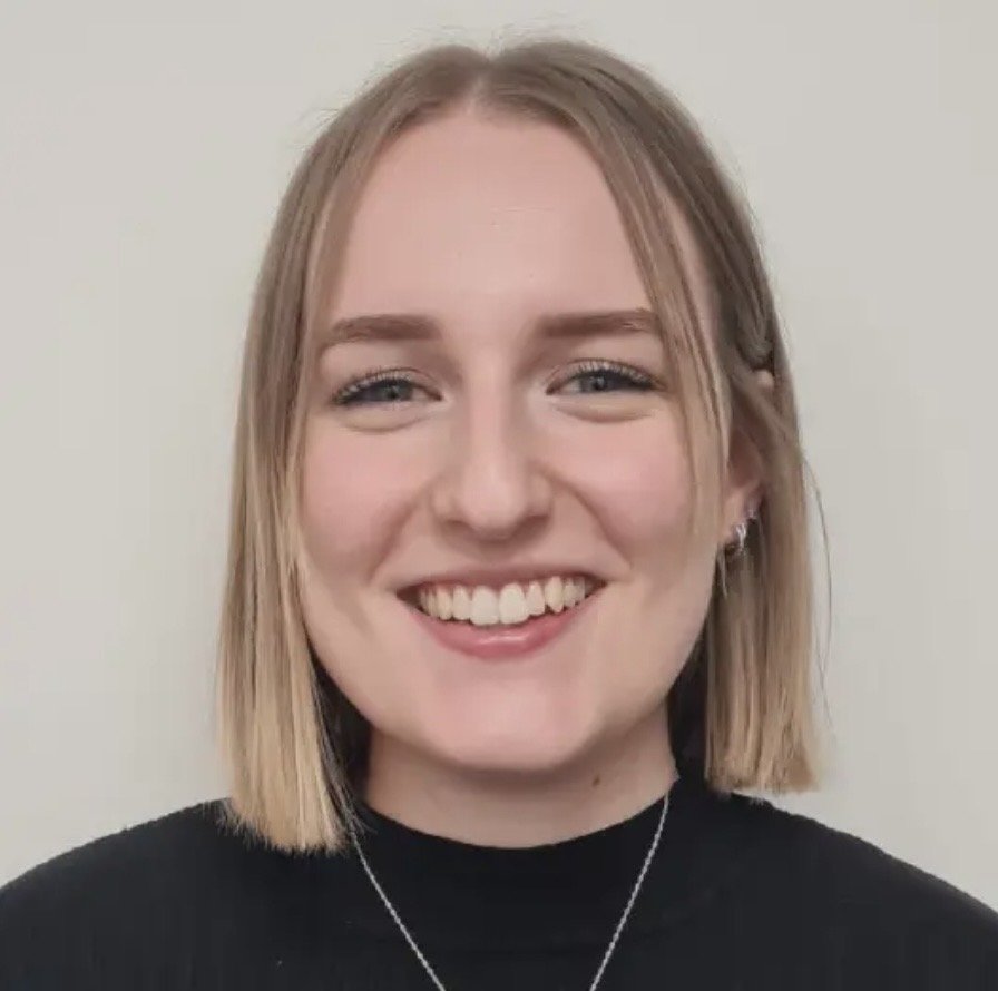 A young woman with blonde, shoulder-length hair, smiling at the camera, wearing a black top and a silver necklace, against a plain light background.
