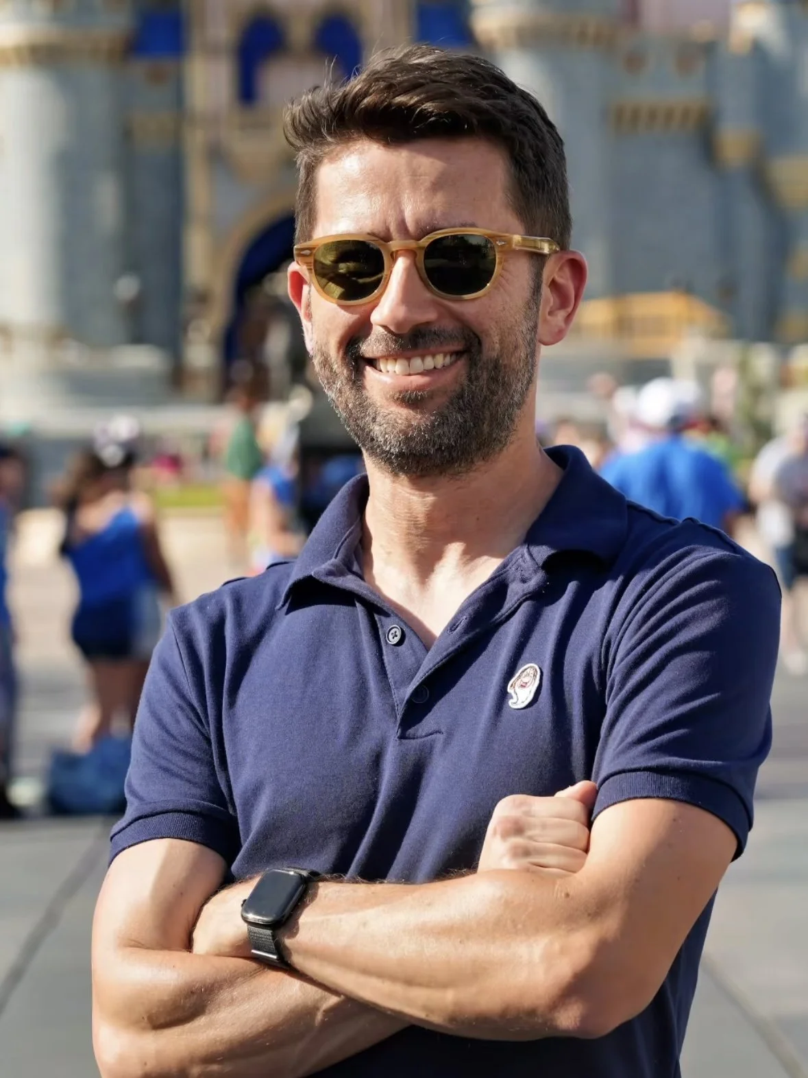 Smiling man wearing a personally styled outfit with sunglasses and a navy polo shirt standing outdoors in front of Cinderella Castle at Walt Disney World, with other visitors in the background.