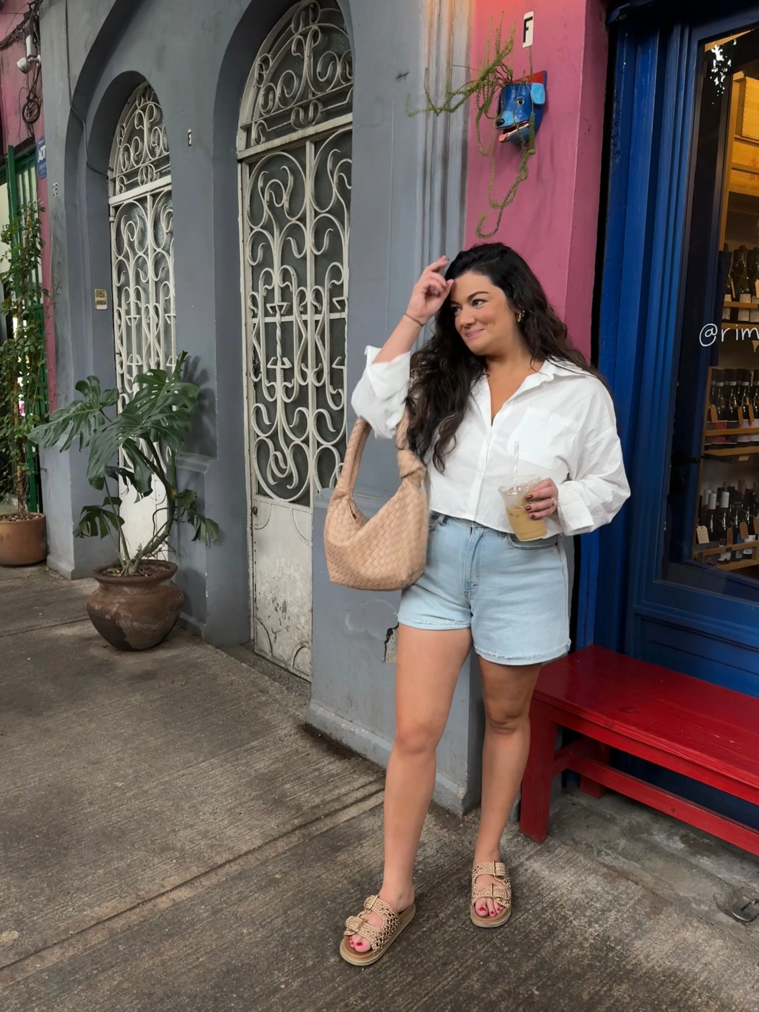 A woman with long dark curly hair wearing a vacation outfit: a white shirt, light denim shorts, and sandals, standing on a sidewalk outside a colorful building, holding a drink, with potted plants and decorative ironwork in the background.